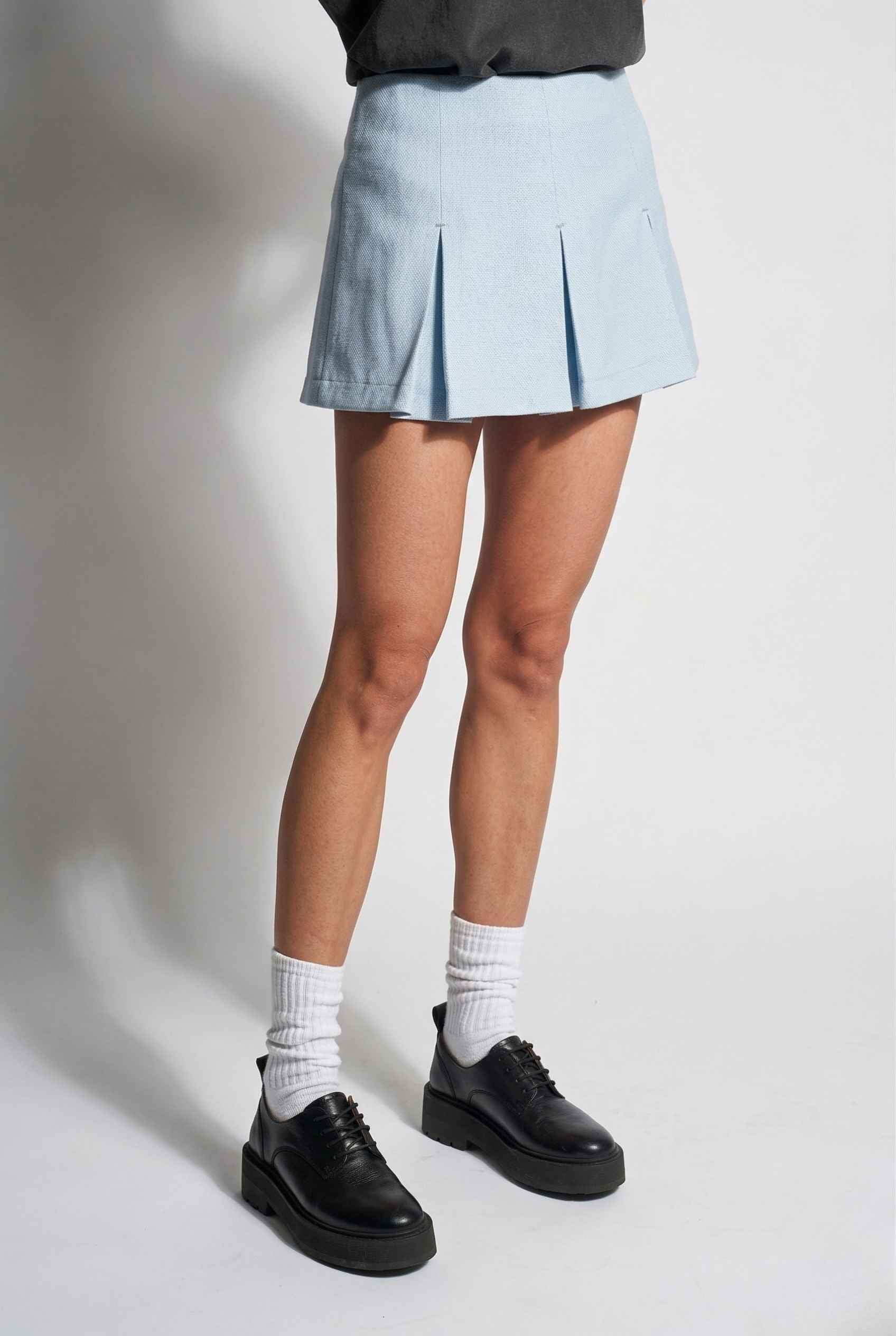 Woman wearing a Celeste Skort Baby Blue Pleated Silk-Blend Mini Skort, black shoes, and white socks on a plain background