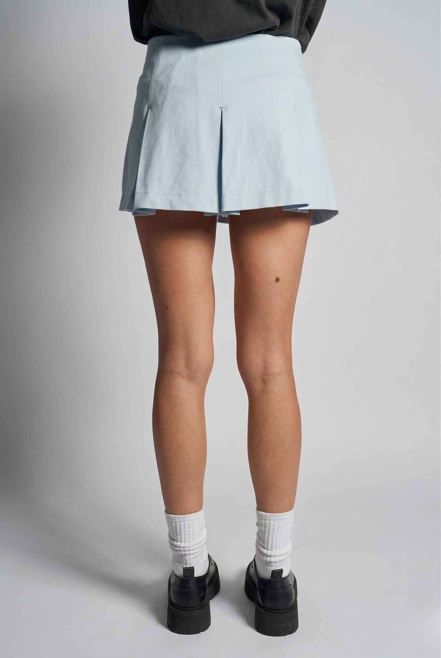 Woman wearing a Celeste Skort Baby Blue Pleated Silk-Blend Mini Skort, white socks, and black shoes on a plain background