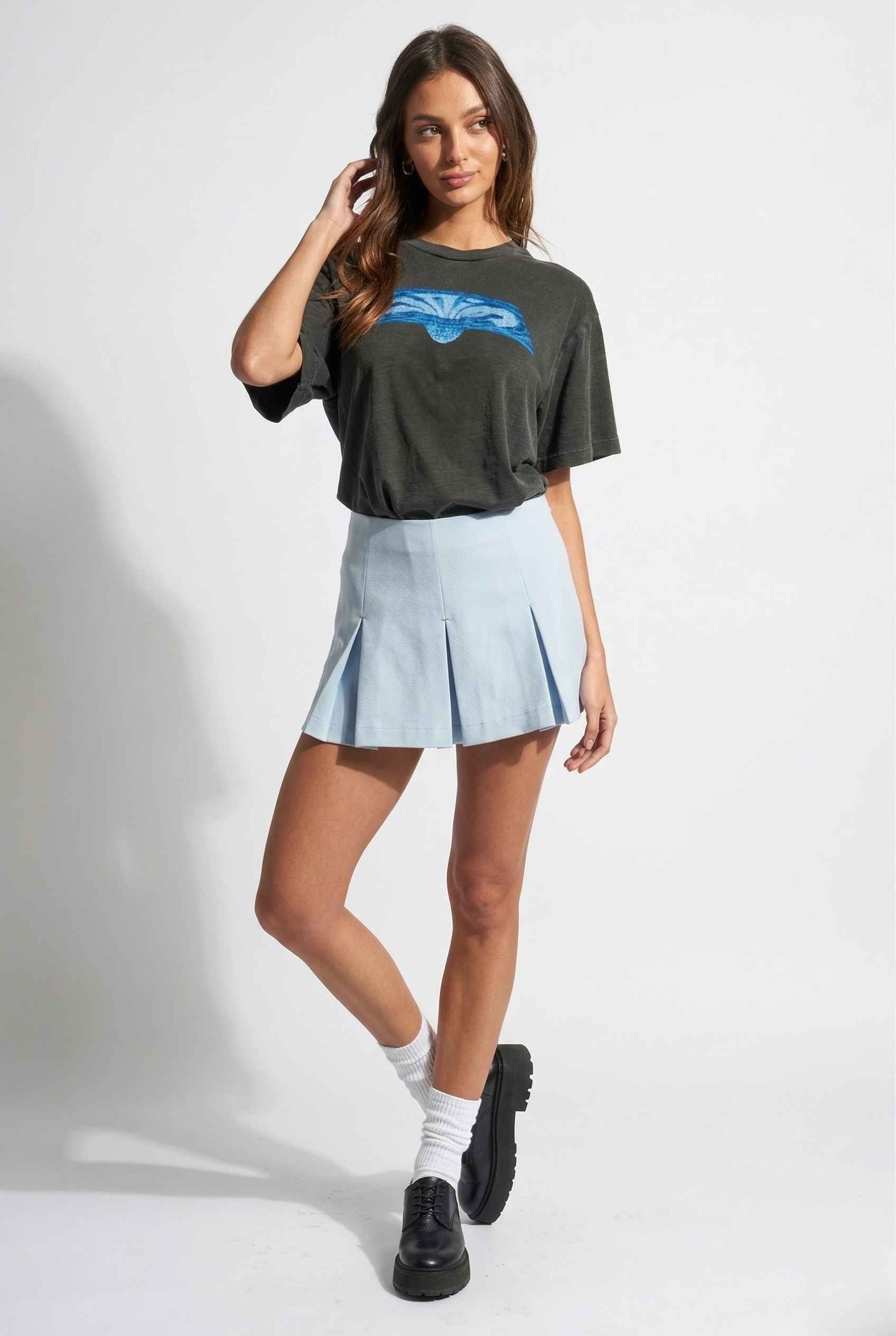 Woman wearing a dark green oversized t-shirt with a blue graphic and a Celeste Skort Baby Blue Pleated Silk-Blend Mini Skort on a white background