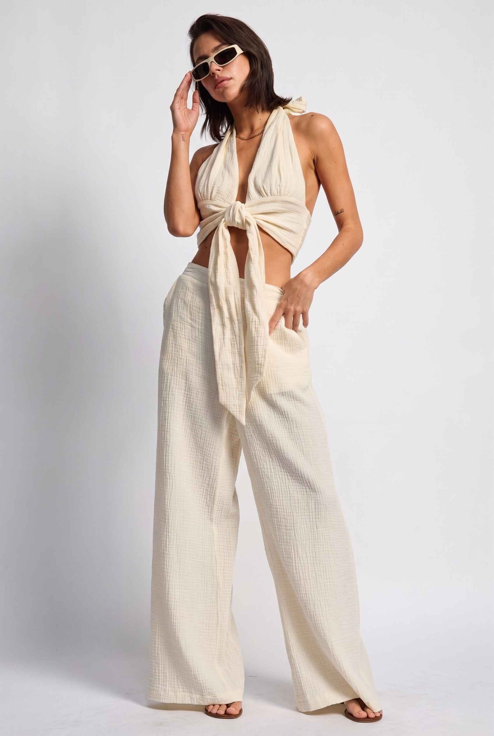 Model wearing Celine Gauze Halter Crop Top 100% Cotton Tie-Front Top in Off White paired with matching high waisted pants and sandals
