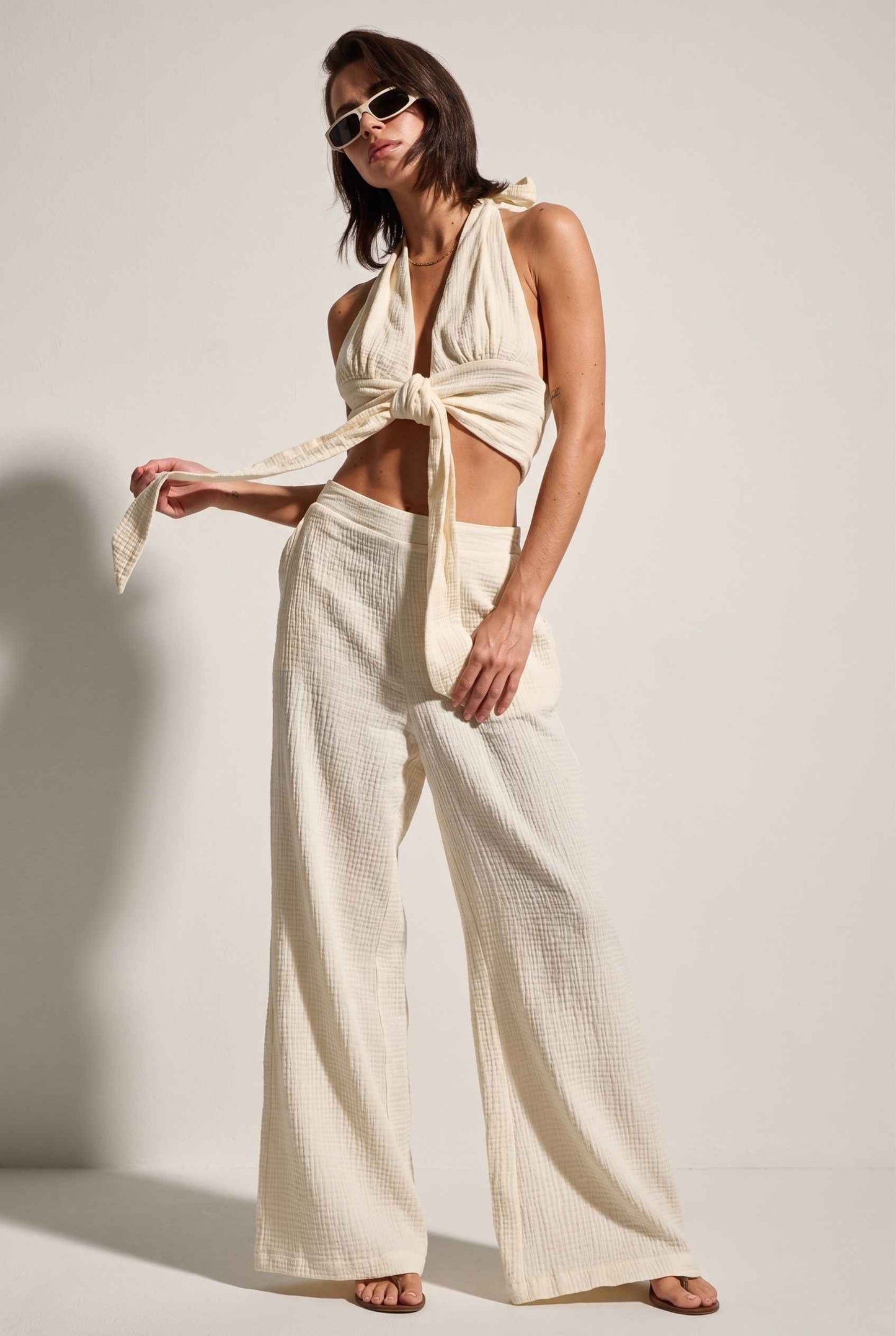Model wearing Celine Gauze Halter Crop Top 100% Cotton Tie-Front Top in Off White paired with matching high waisted pants and sandals