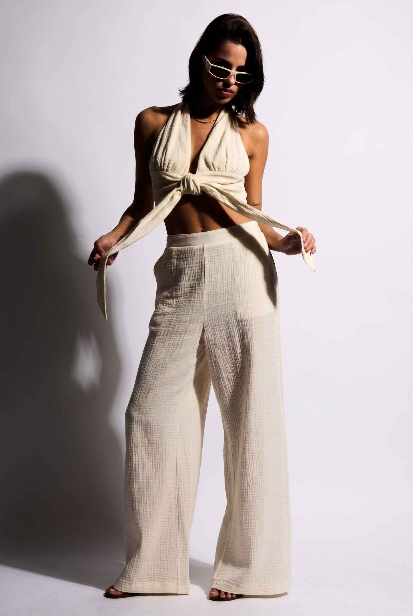 Model wearing Celine Gauze Halter Crop Top 100% Cotton Tie-Front Top in Off White paired with matching high waisted pants and sandals