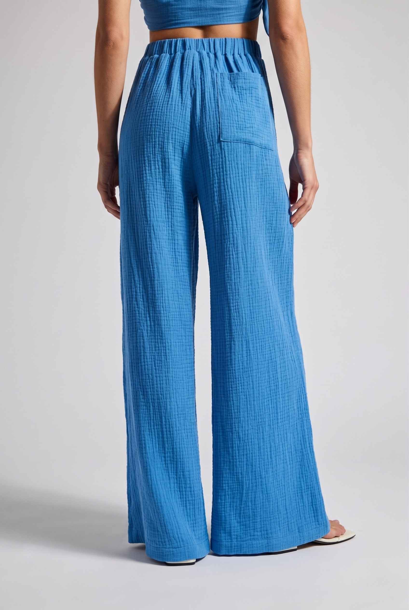 Model wearing Celine High-Waisted Gauze Pant 100% Cotton Wide-Leg Resort Pant in Blue paired with matching halter crop top and sandals close back view