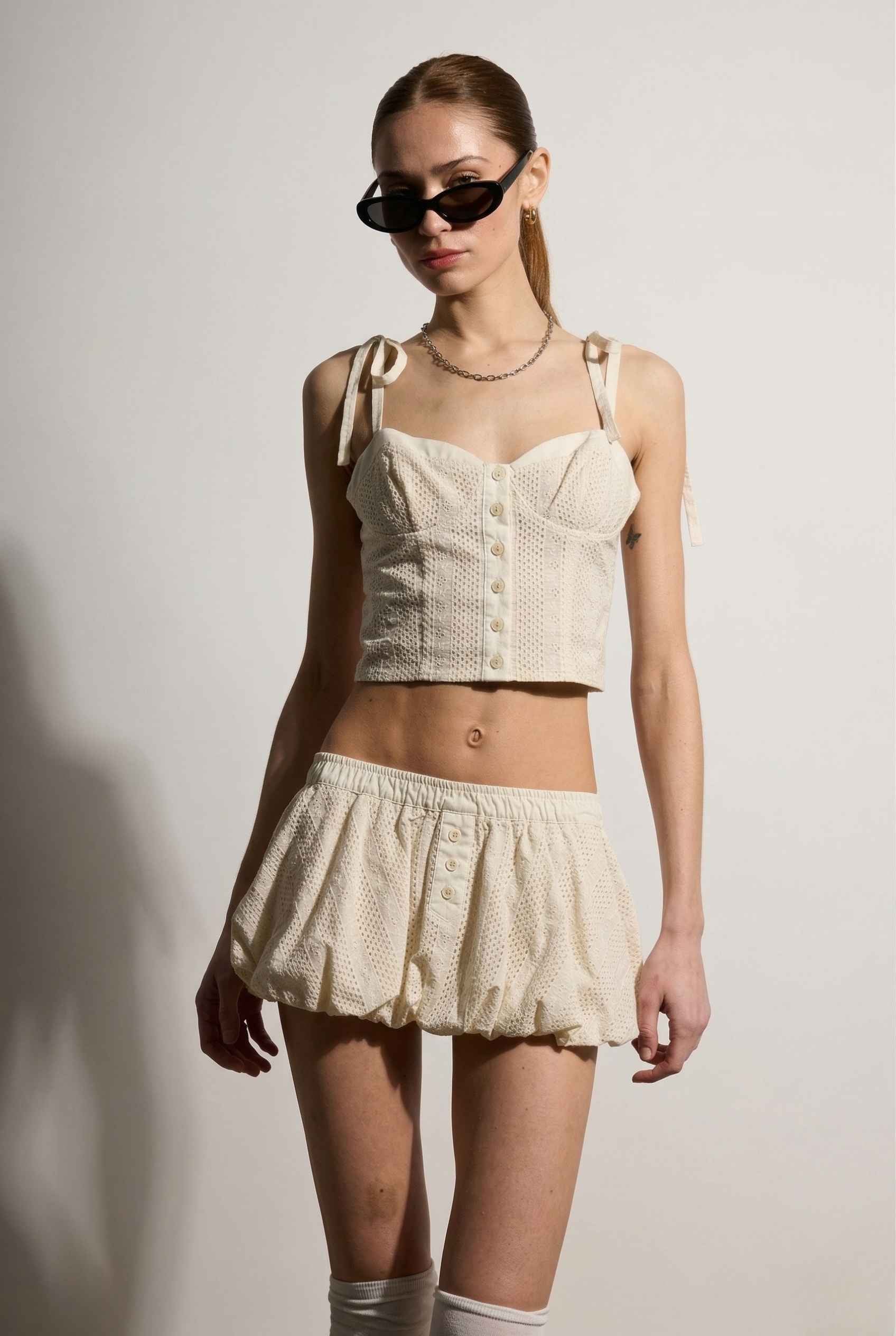 Woman wearing a cream-colored top and Chloe Skirt Cream Cotton Eyelet Bubble Hem Mini Skirt with sunglasses on a plain background