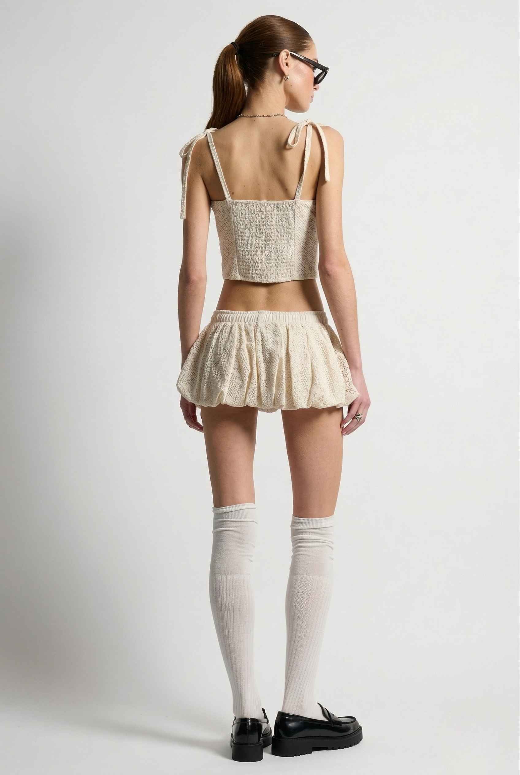 Woman wearing a white sleeveless top and Chloe Skirt Cream Cotton Eyelet Bubble Hem Mini Skirt with knee-high socks on a plain background