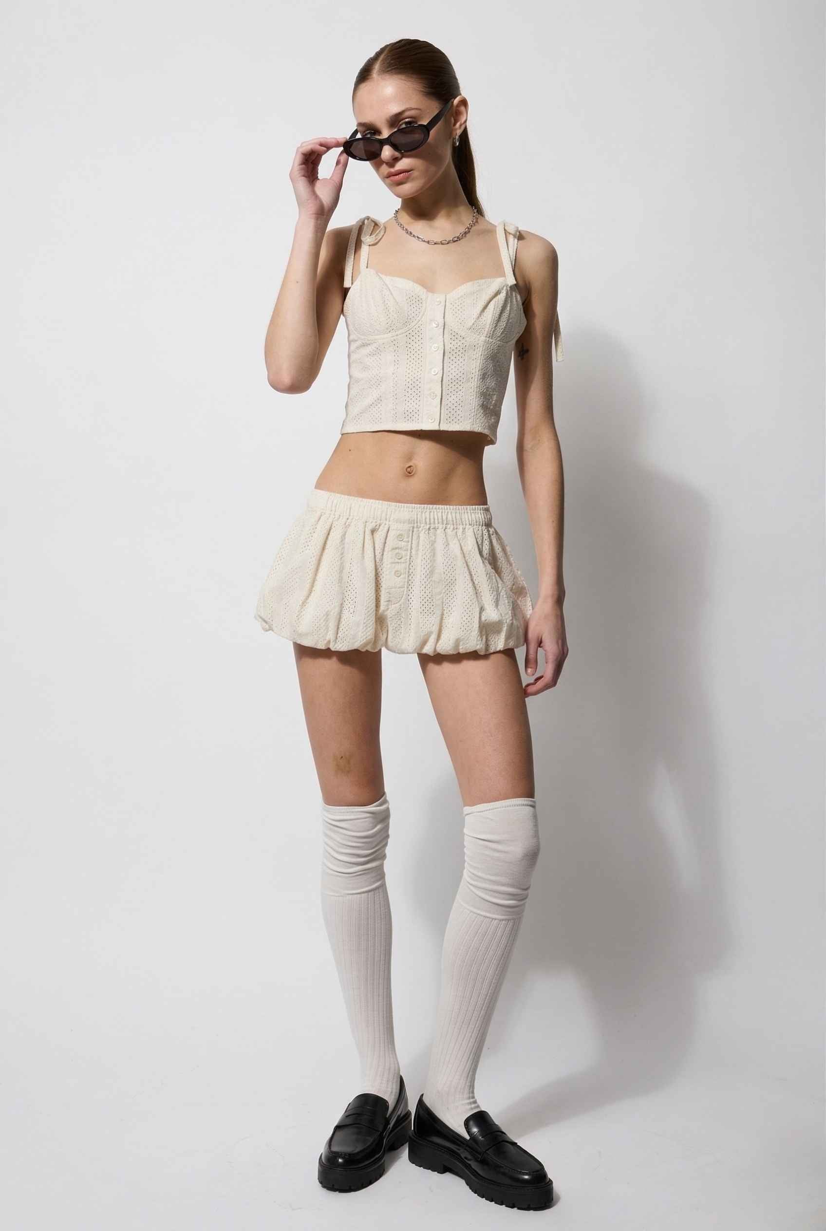 Woman wearing a beige top and Chloe Skirt Cream Cotton Eyelet Bubble Hem Mini Skirt with white stockings and black shoes on a white background