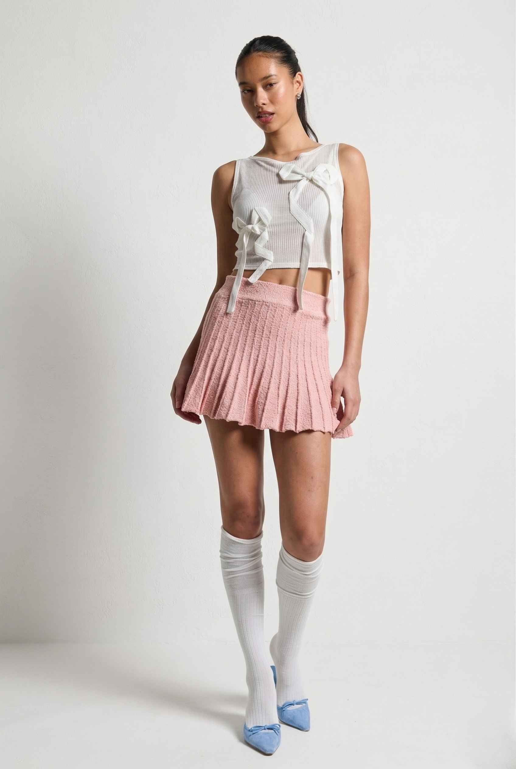 Woman wearing a sleeveless top with a bow, Chloe Skirt Textured Knit Pleated Mini Skirt in Pink, and knee-high socks on a background