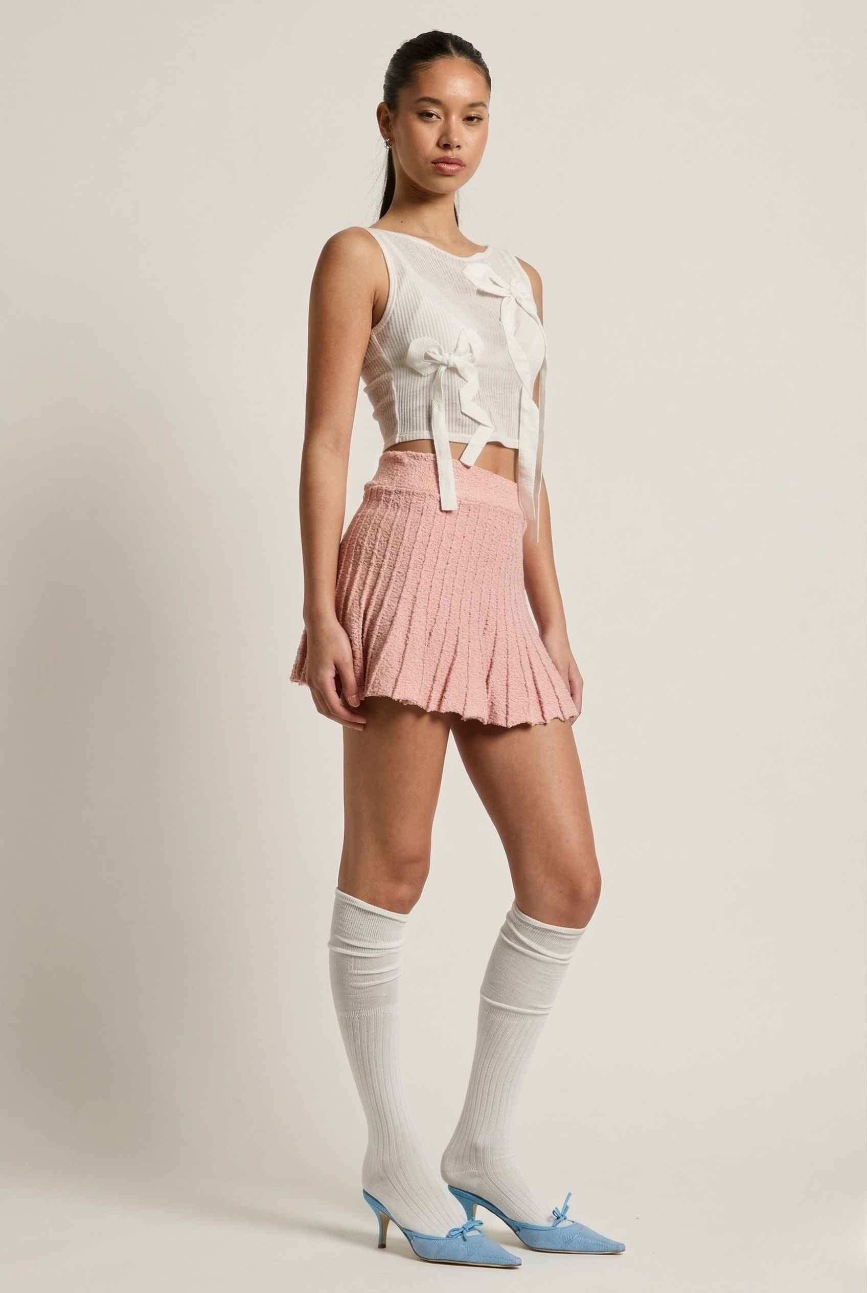 Woman wearing a sleeveless top, Chloe Skirt Textured Knit Pleated Mini Skirt in Pink, and knee-high socks with blue shoes on a plain background