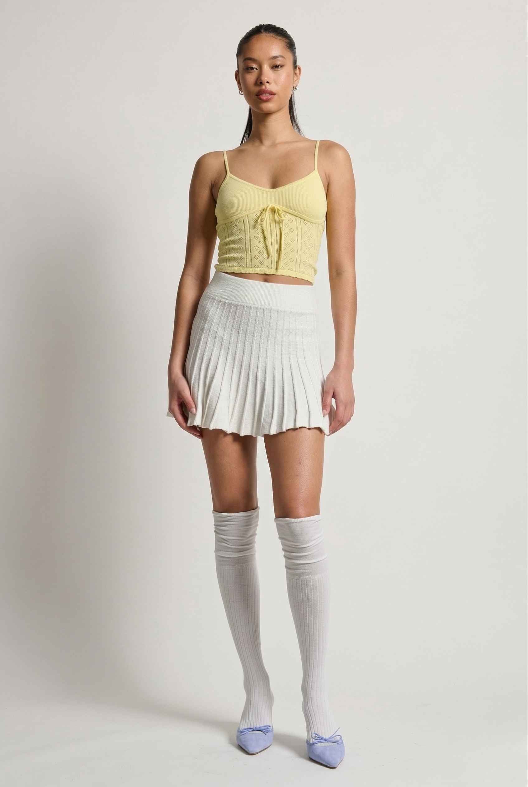 Woman wearing a yellow top, Chloe Skirt Textured Knit Pleated Mini Skirt in White, and knee-high socks on a plain background