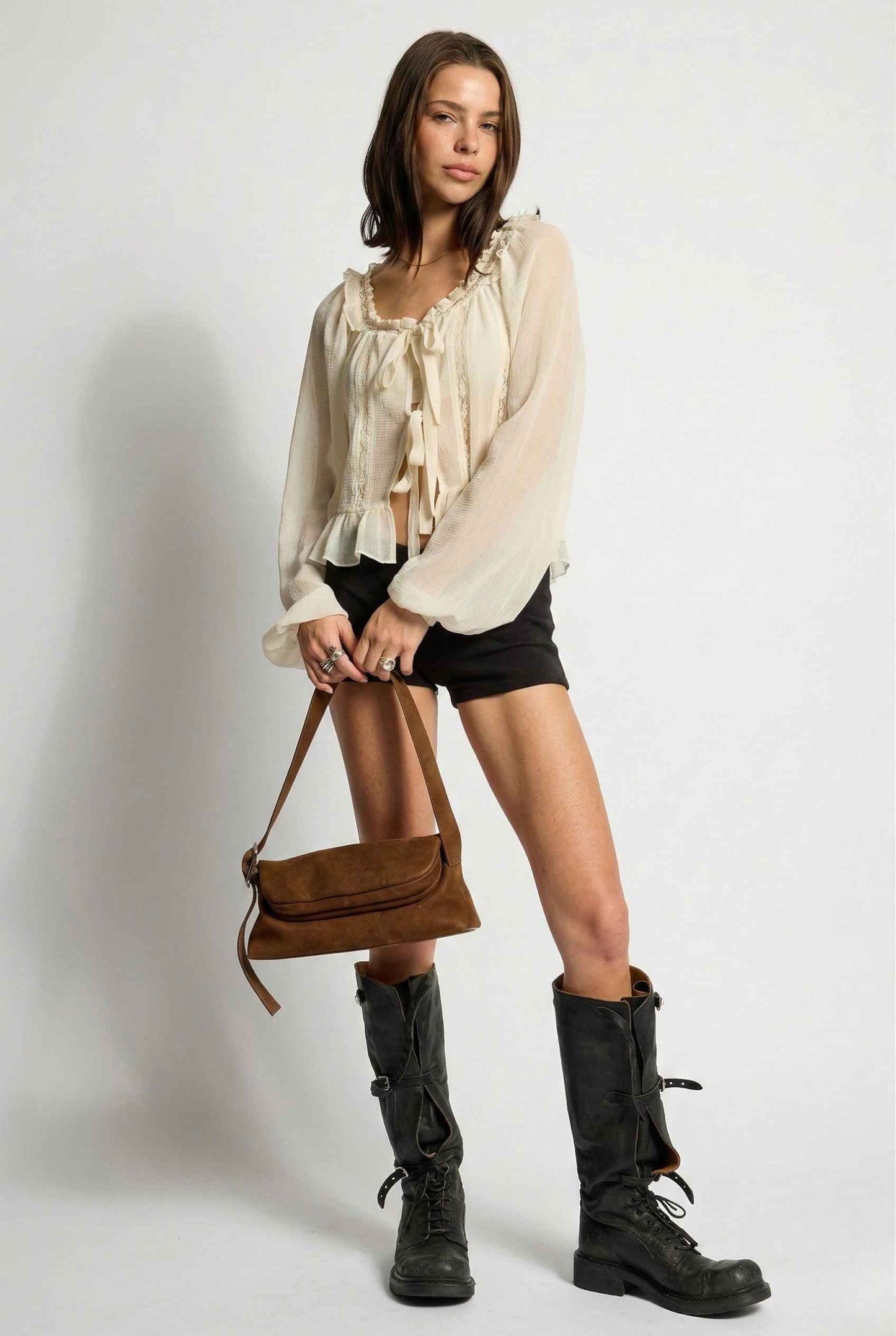 Woman wearing a Clara Top Cream Sheer Chiffon Tie-Front Balloon Sleeve Blouse, black shorts, and black boots holding a brown handbag against a white background