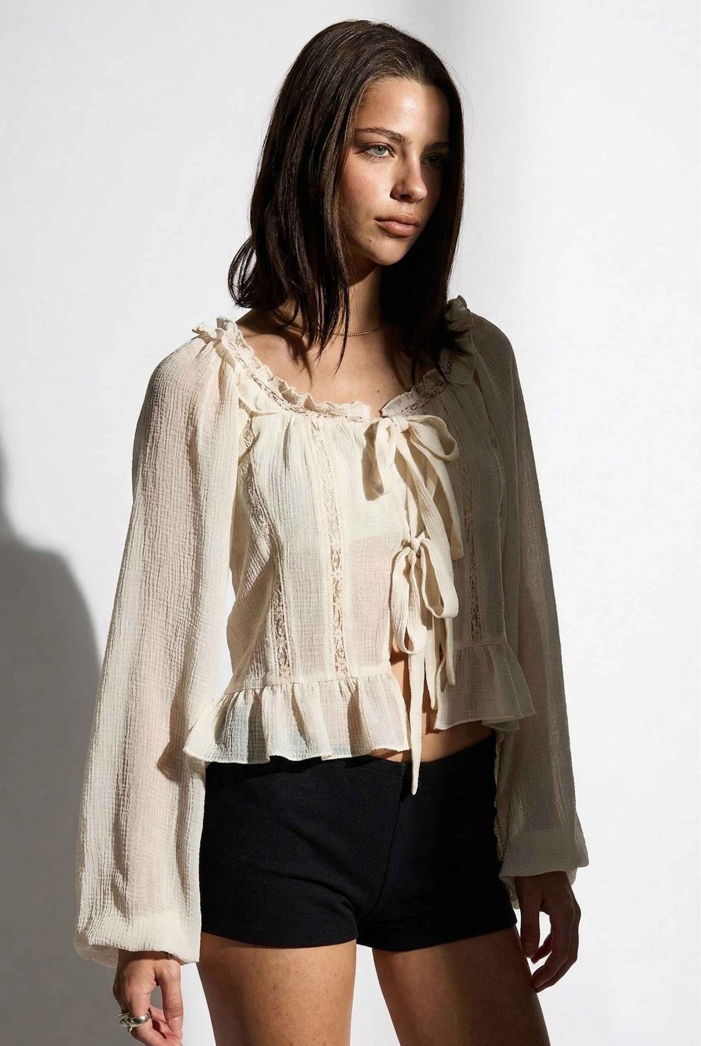 Woman wearing a Clara Top Cream Sheer Chiffon Tie-Front Balloon Sleeve Blouse with long sleeves and black shorts on a white background