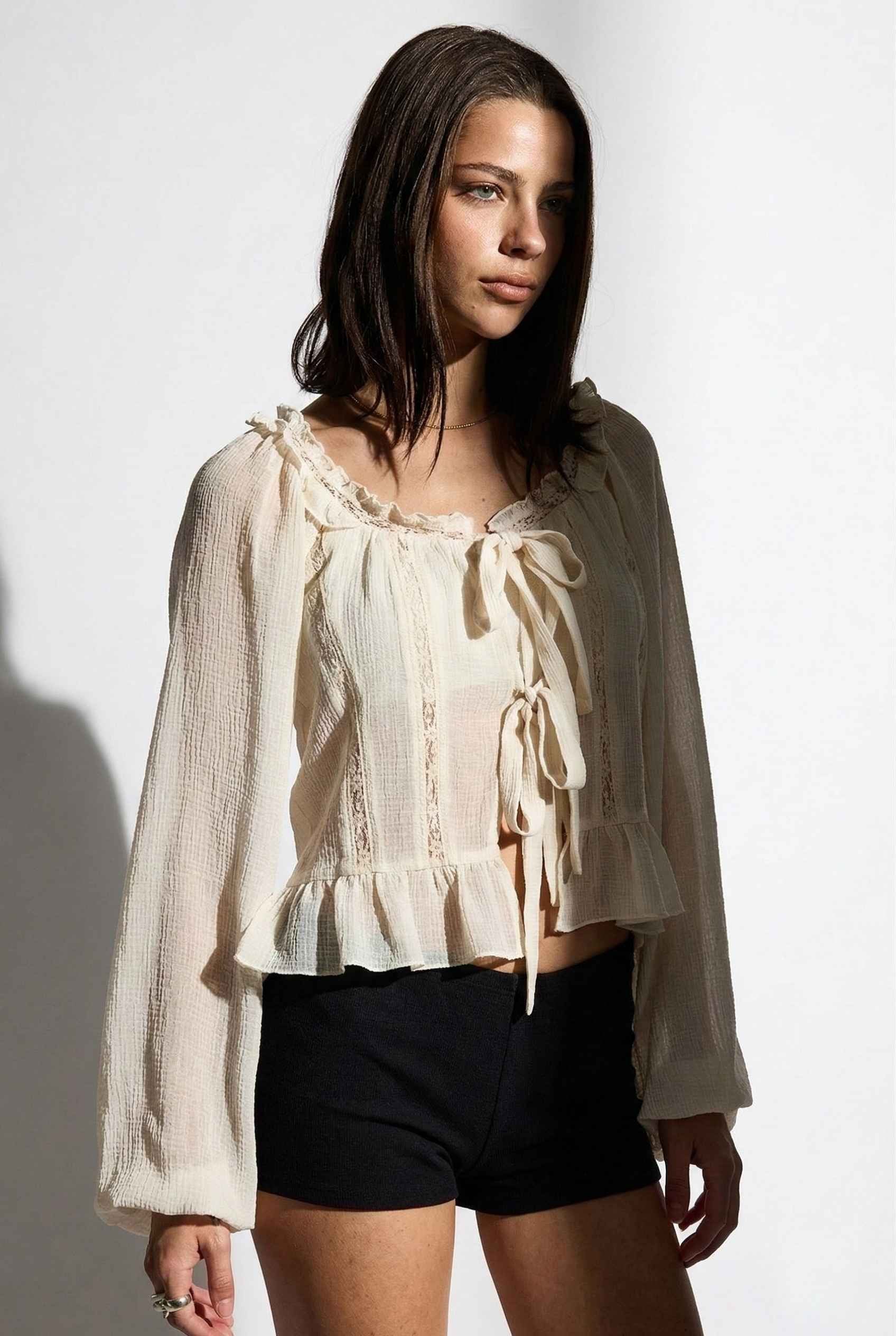 Woman wearing a Clara Top Cream Sheer Chiffon Tie-Front Balloon Sleeve Blouse with long sleeves and black shorts on a white background