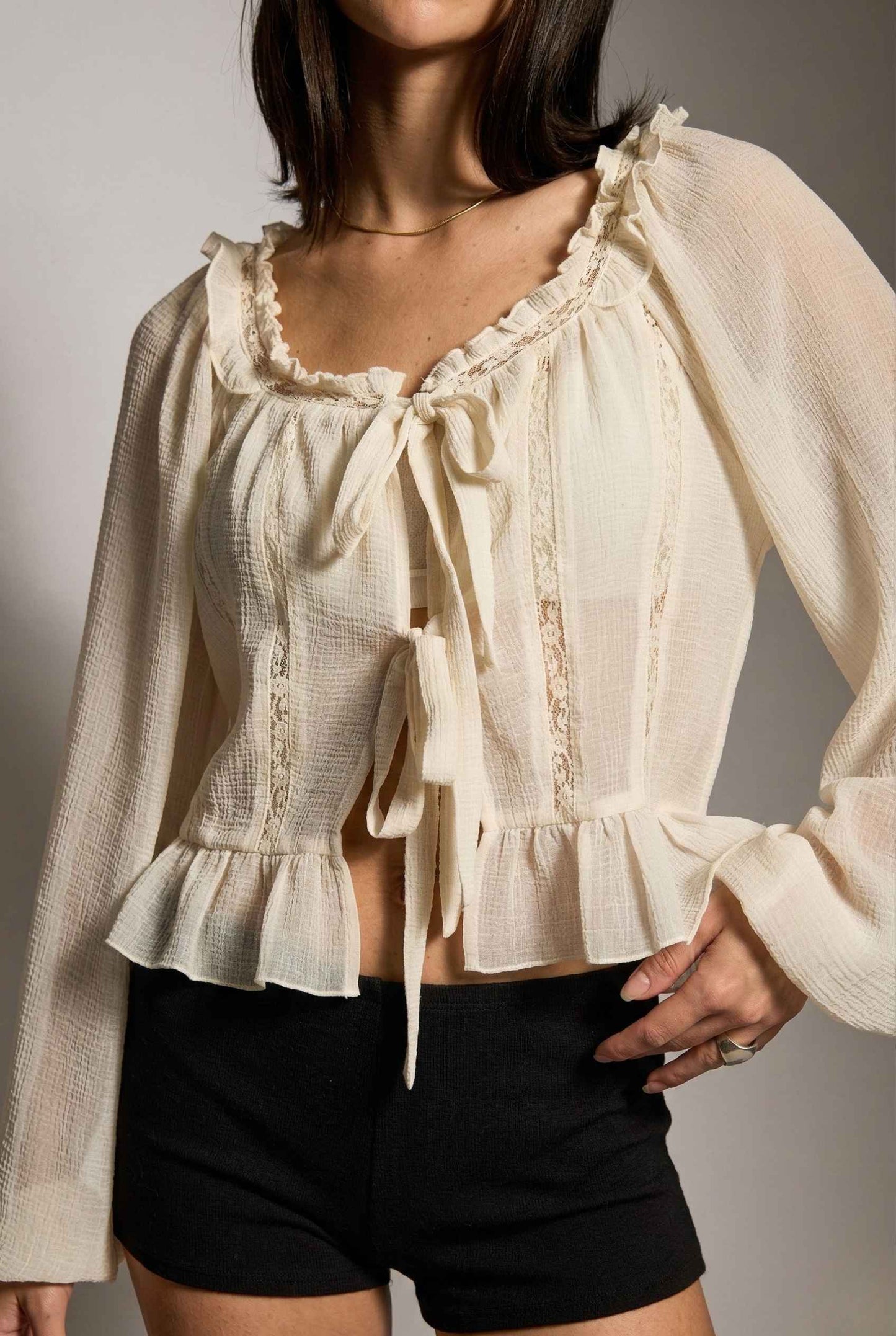 Woman wearing a Clara Top Cream Sheer Chiffon Tie-Front Balloon Sleeve Blouse with lace details and black shorts against a neutral background