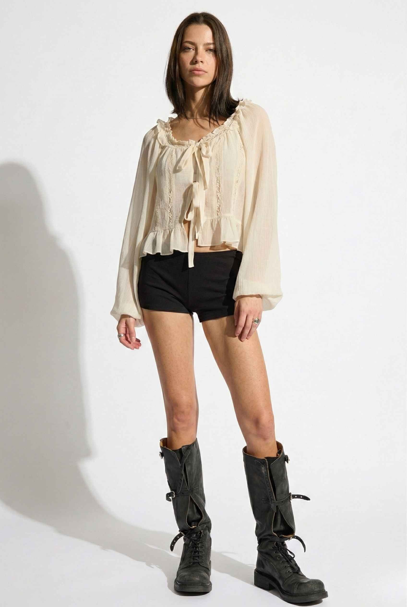 Woman wearing a Clara Top Cream Sheer Chiffon Tie-Front Balloon Sleeve Blouse, black shorts, and black boots on a white background
