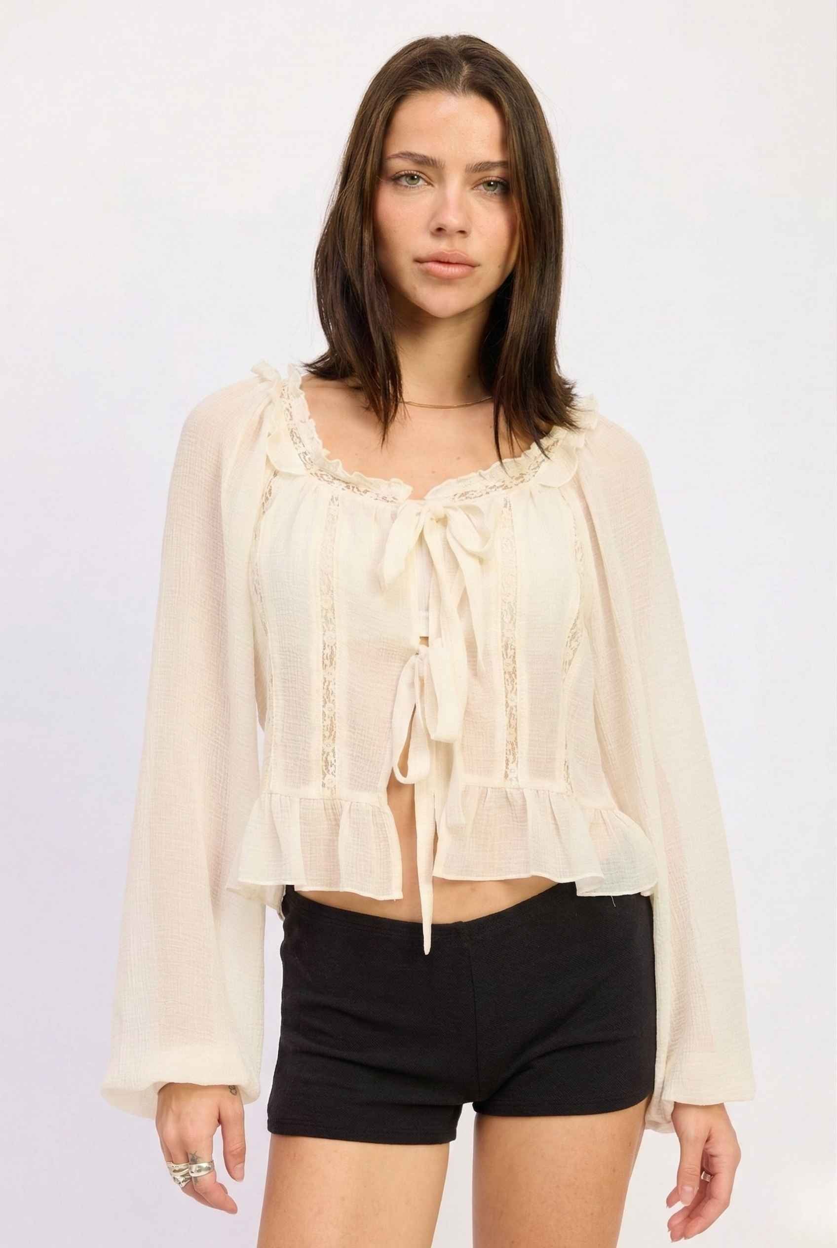 Woman wearing a Clara Top Cream Sheer Chiffon Tie-Front Balloon Sleeve Blouse with ruffles and black shorts on a white background