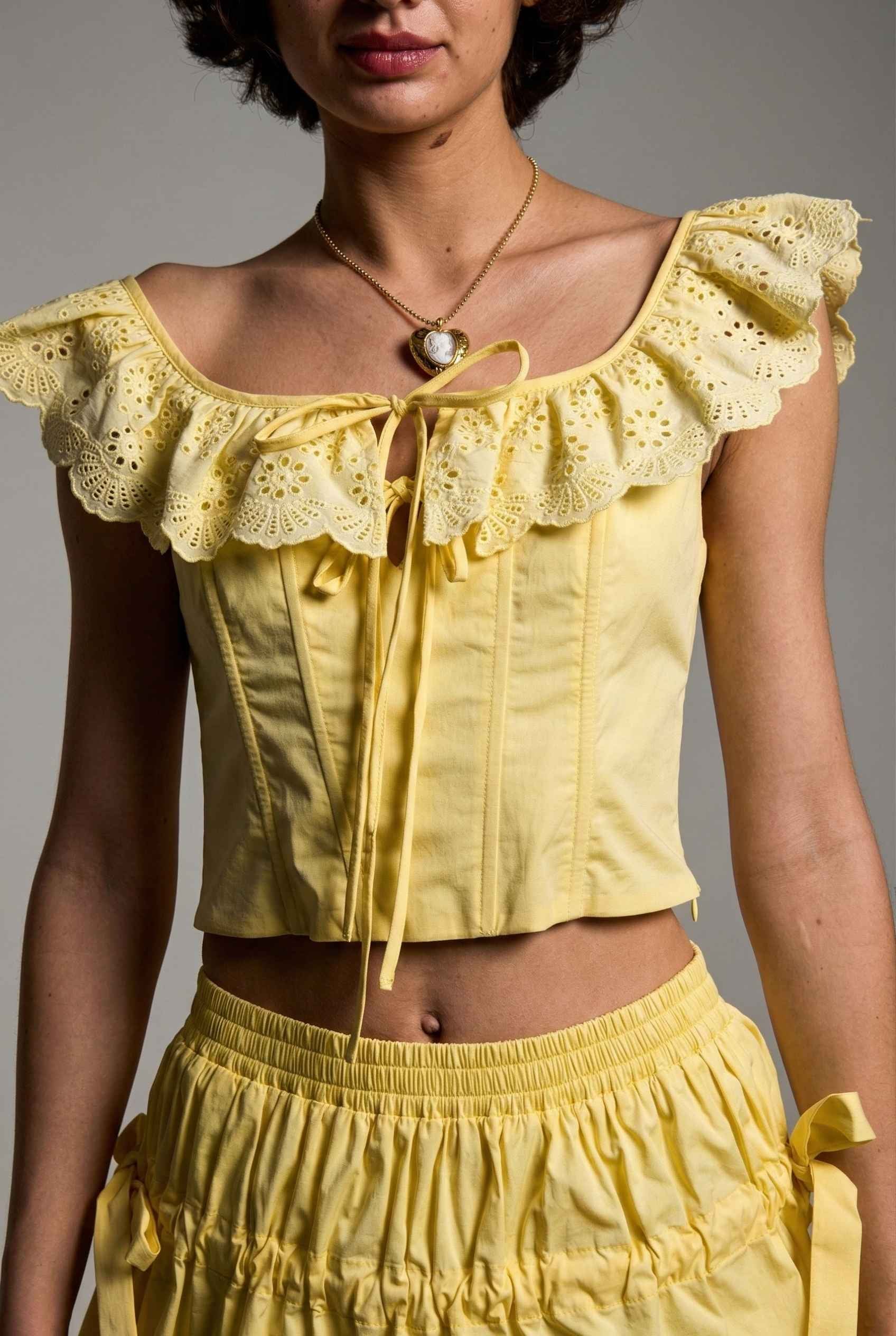 Woman wearing a Clementine Top Lemon Yellow Cotton Eyelet Ruffle Corset and skirt against a gray background