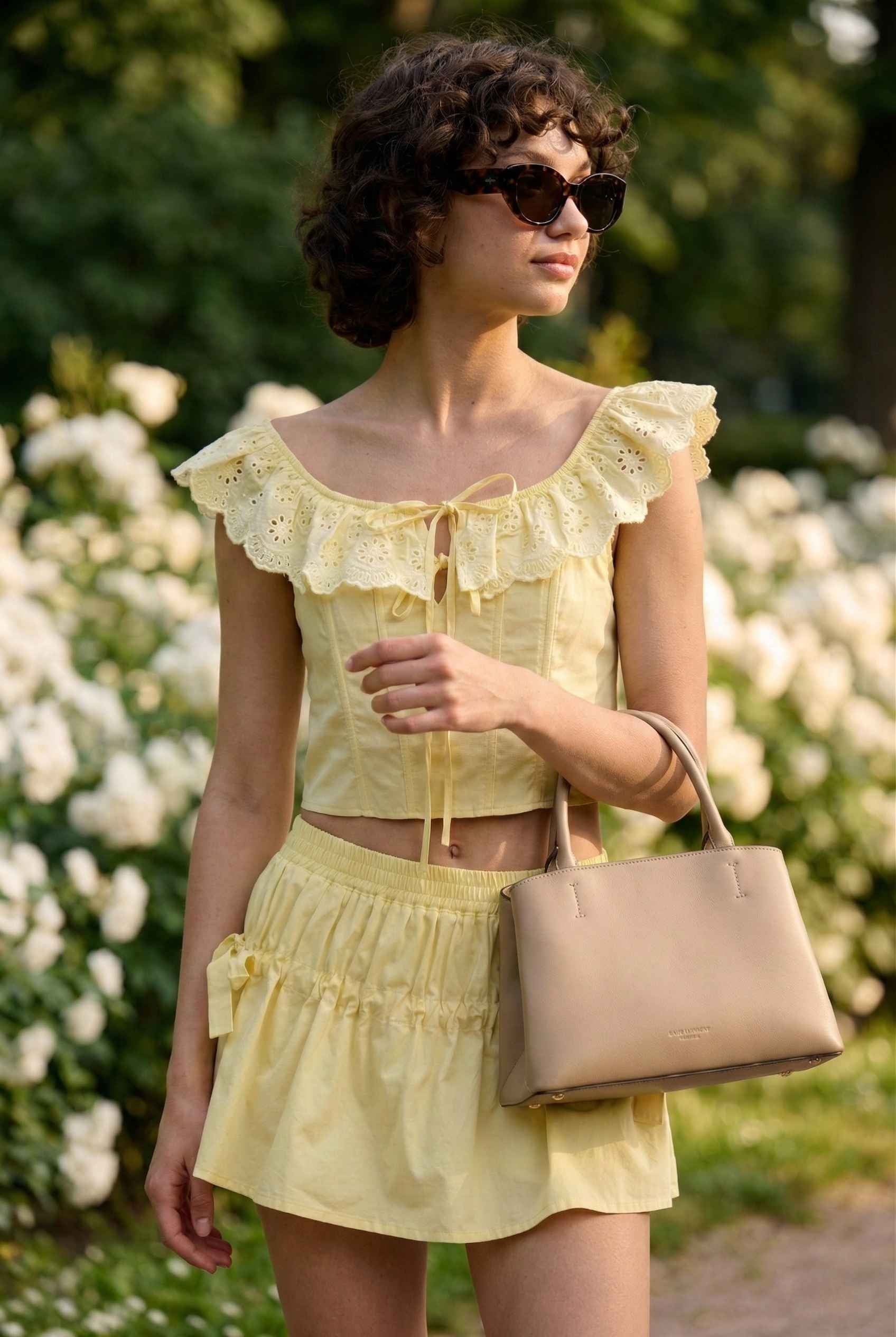 Woman in a Clementine Top Lemon Yellow Cotton Eyelet Ruffle Corset holding a beige handbag in a garden setting