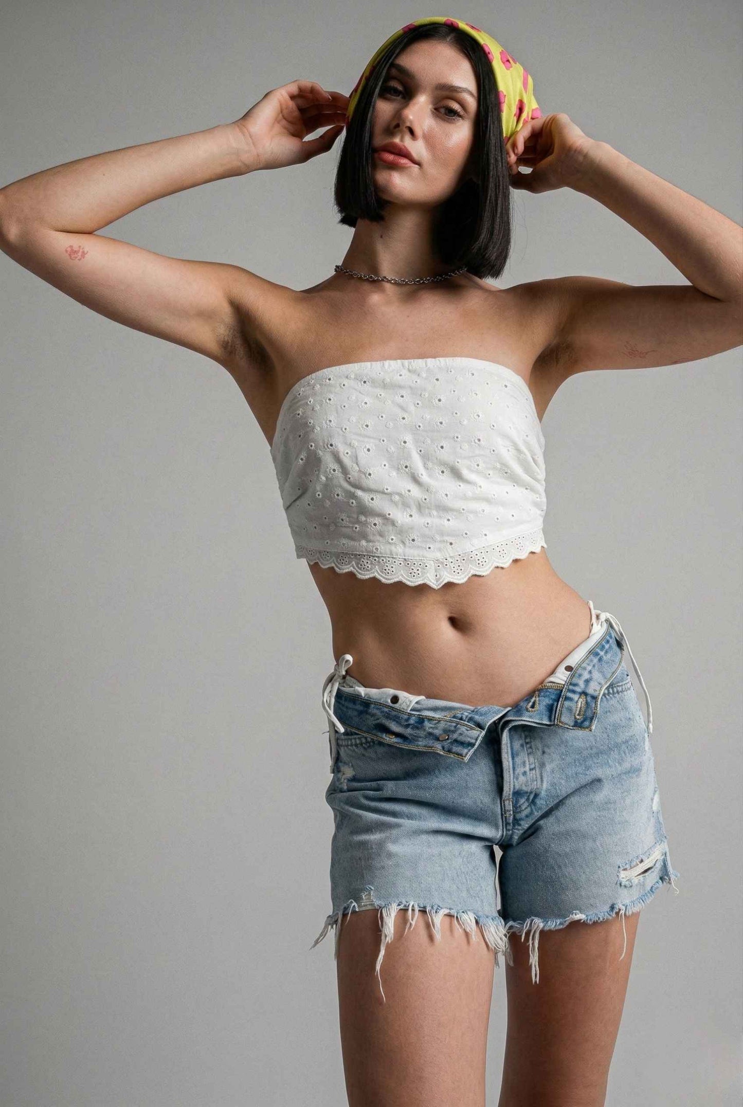 Woman wearing a Clementine Top White Cotton Eyelet Strapless Crop and denim shorts against a gray background