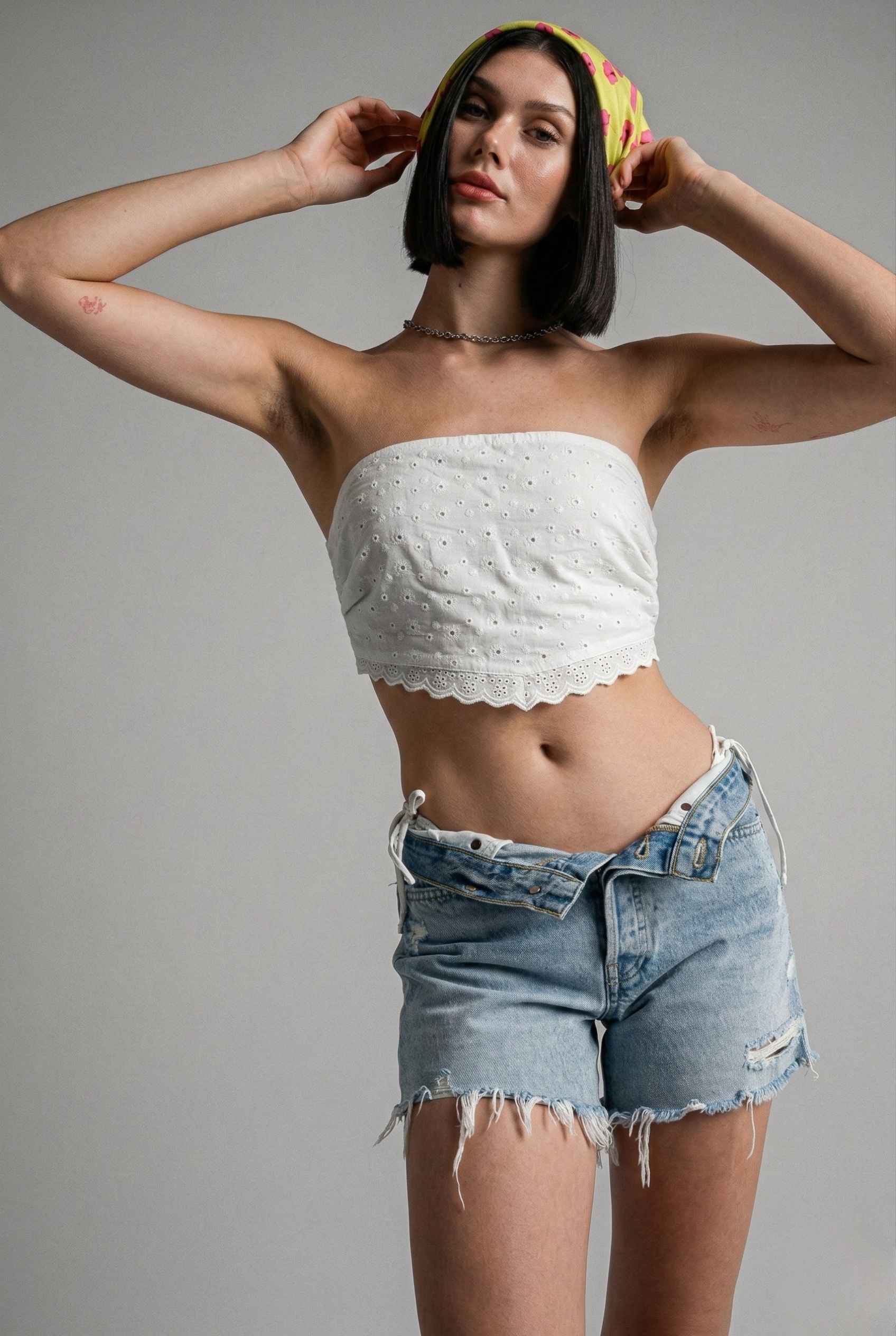 Woman wearing a Clementine Top White Cotton Eyelet Strapless Crop and denim shorts against a gray background