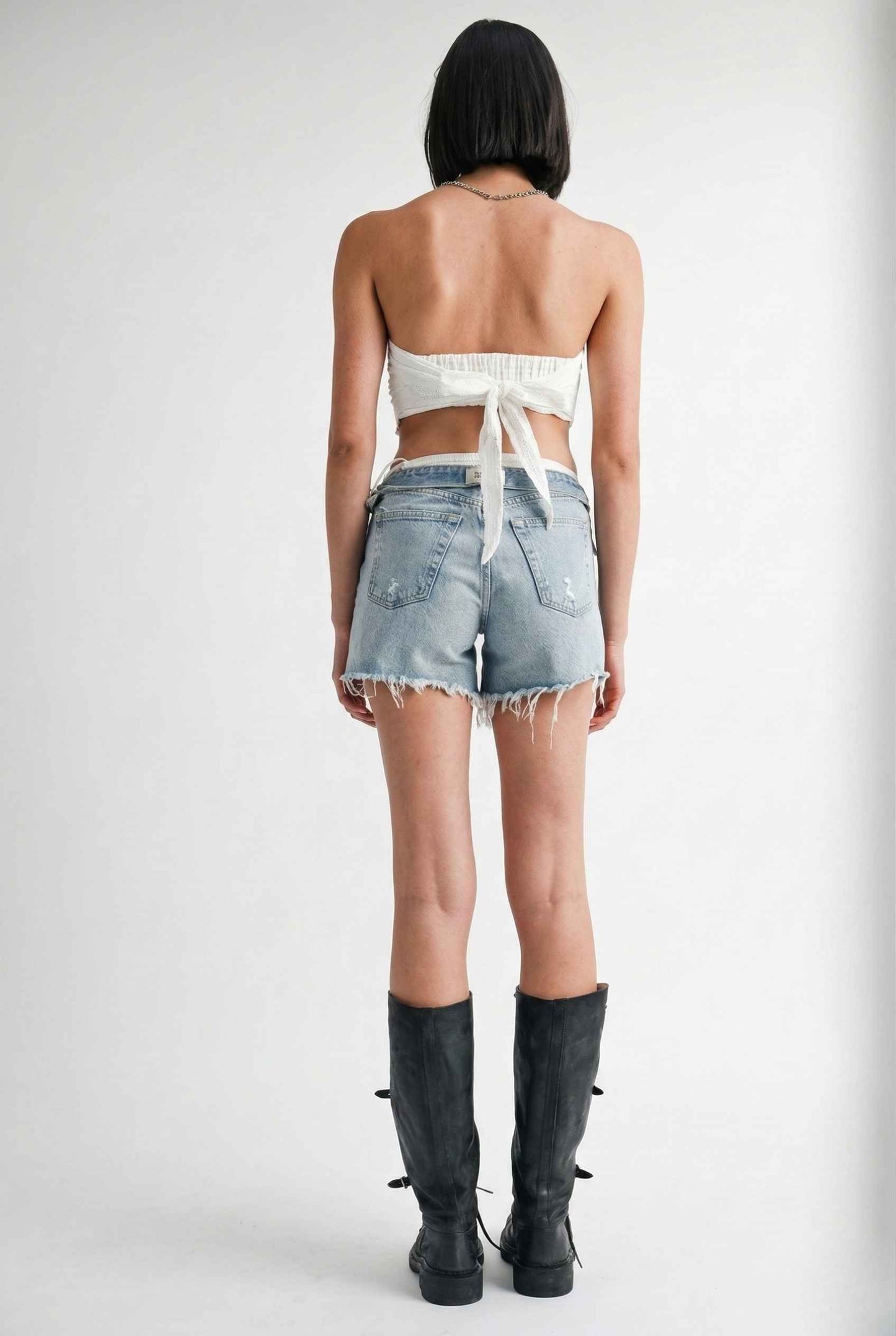 Woman wearing a Clementine Top White Cotton Eyelet Strapless Crop and denim shorts with black boots on a plain background