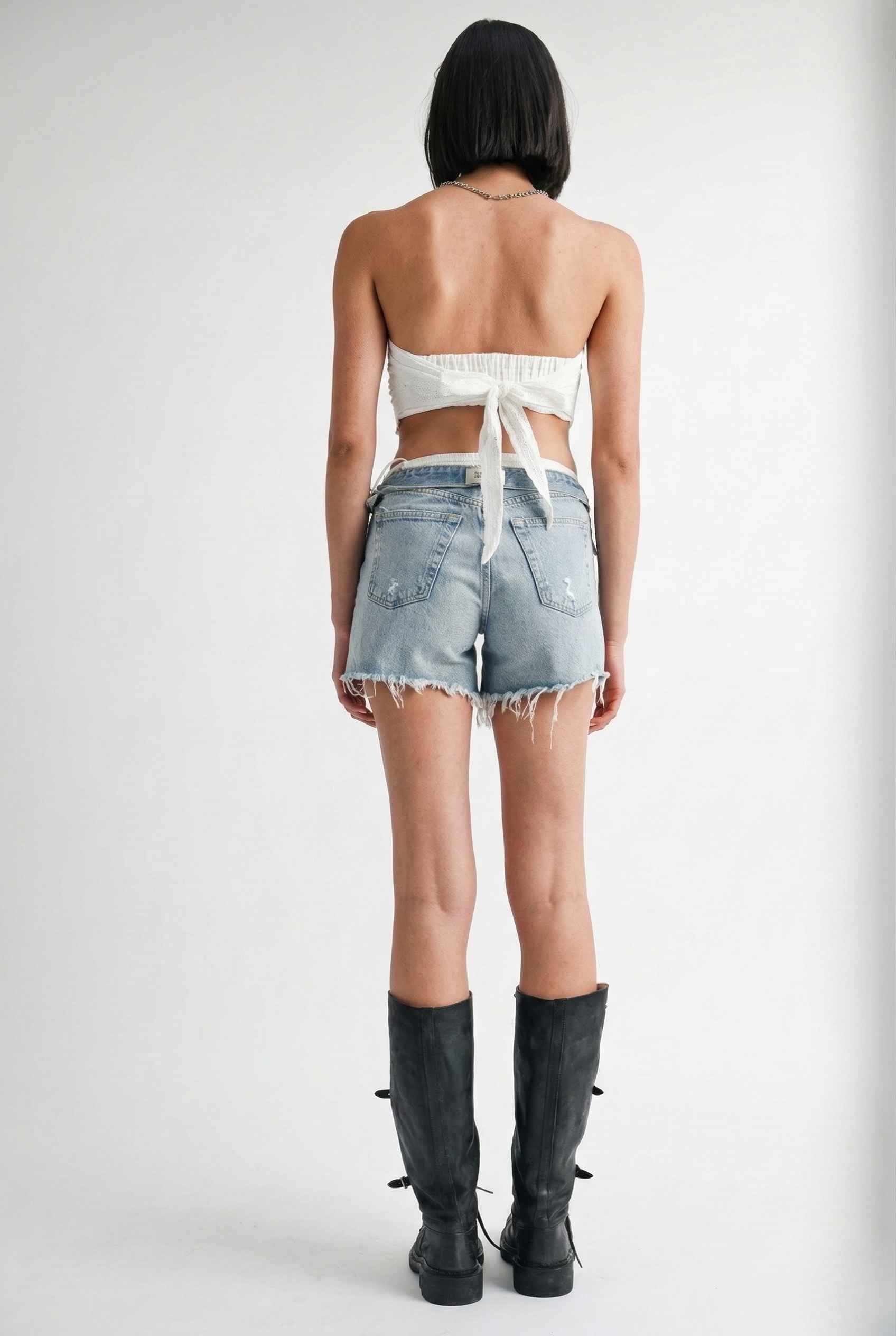 Woman wearing a Clementine Top White Cotton Eyelet Strapless Crop and denim shorts with black boots on a plain background
