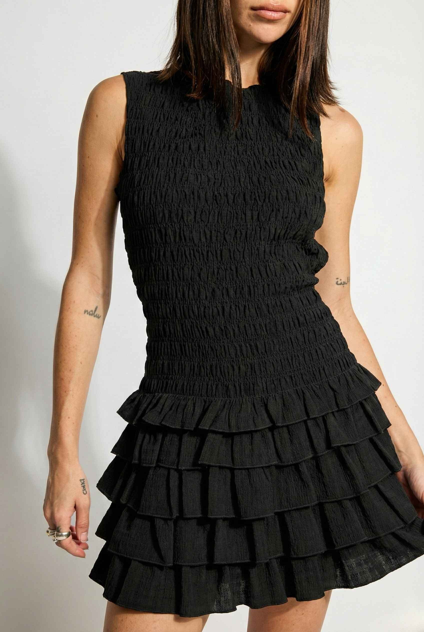 Cleo Mini Black Smocked Ruffle Sleeveless Dress worn by a woman on a plain background