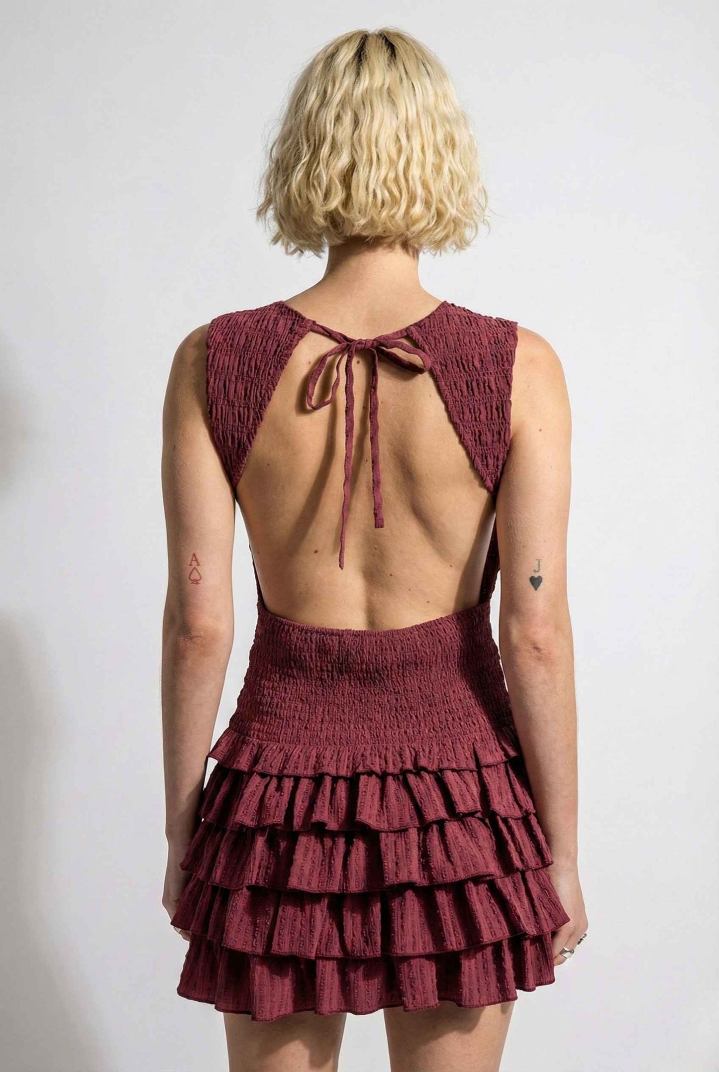 Woman wearing a Cleo Mini Wine Smocked Ruffle Sleeveless Dress with an open back and ruffled layers on a white background