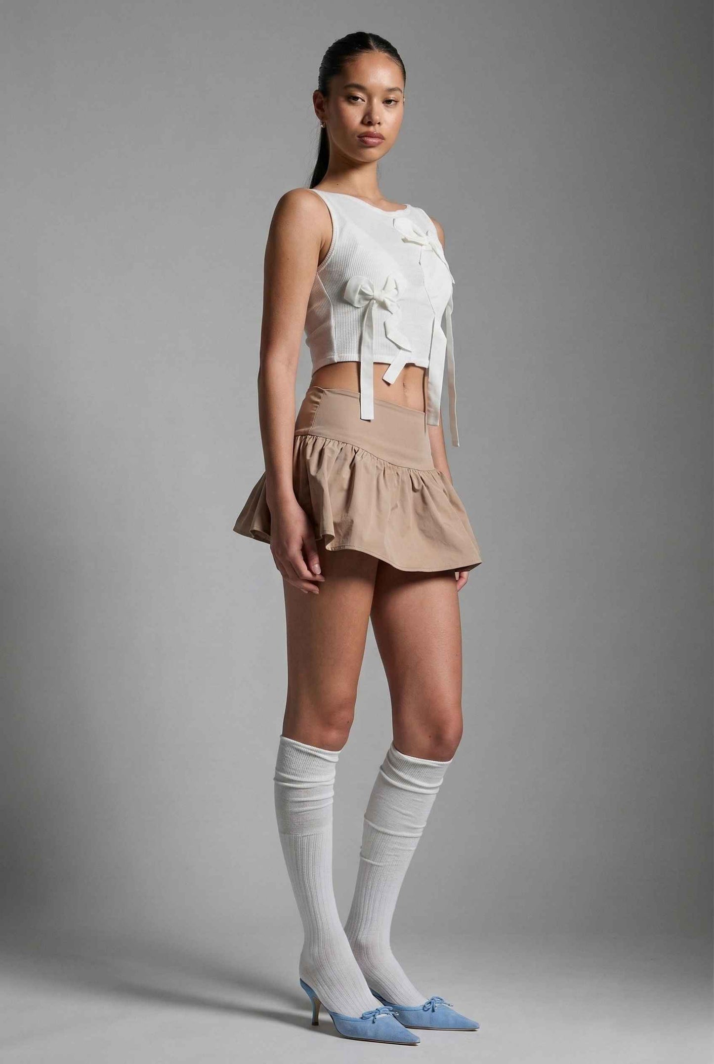 Woman wearing a Colette Top White Sheer Mesh Ribbon Bow Tank and beige skirt with knee-high socks and blue shoes on a gray background