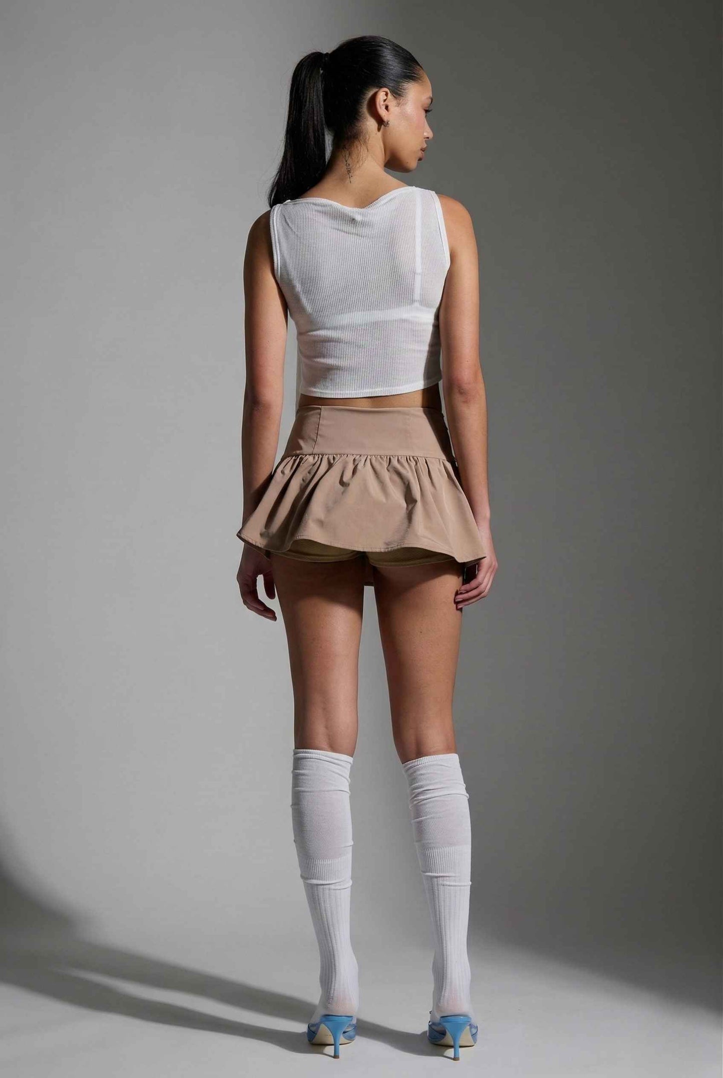 Woman wearing a Colette Top White Sheer Mesh Ribbon Bow Tank and beige skirt with knee-high socks against a gray background