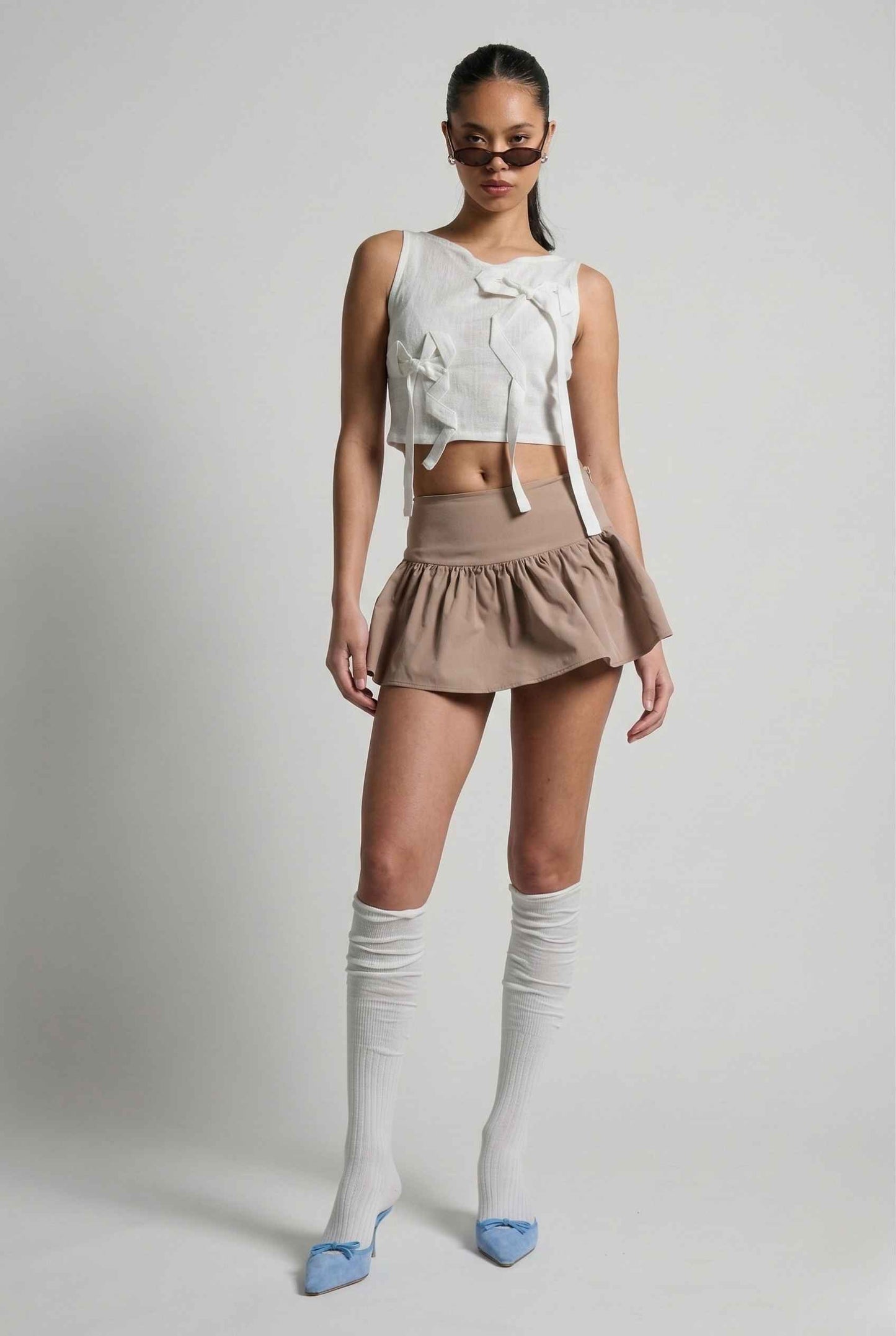 Woman wearing a Colette Top White Sheer Mesh Ribbon Bow Tank, beige skirt, and knee-high socks on a plain background