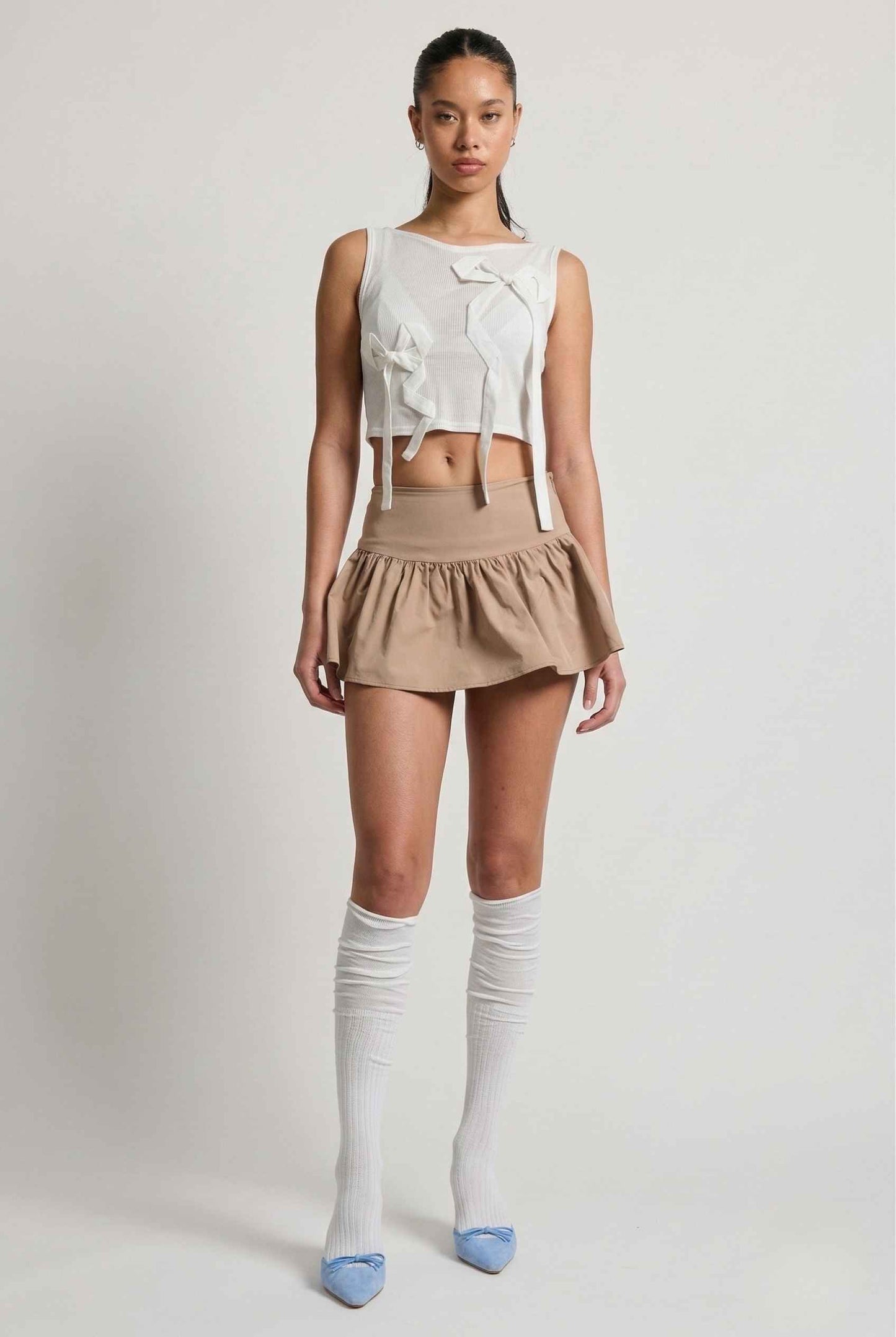 Woman wearing a Colette Top White Sheer Mesh Ribbon Bow Tank with a bow, beige skirt, and knee-high socks on a plain background
