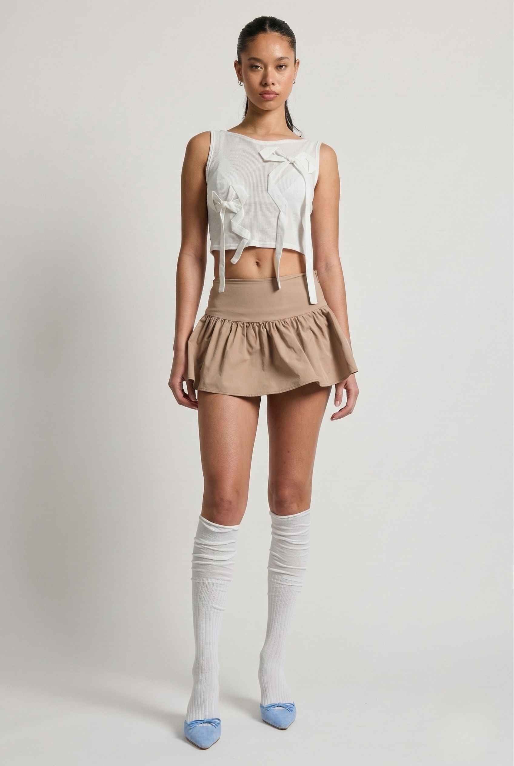 Woman wearing a Colette Top White Sheer Mesh Ribbon Bow Tank with a bow, beige skirt, and knee-high socks on a plain background