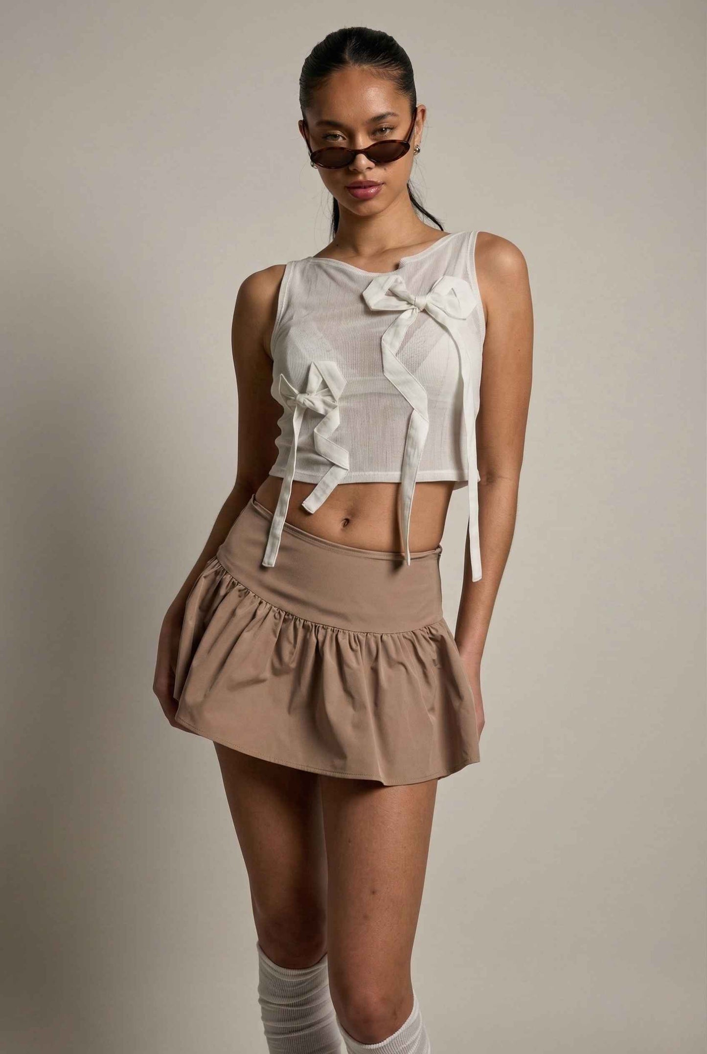 Woman wearing a Colette Top White Sheer Mesh Ribbon Bow Tank with bow details and a beige skirt against a plain background
