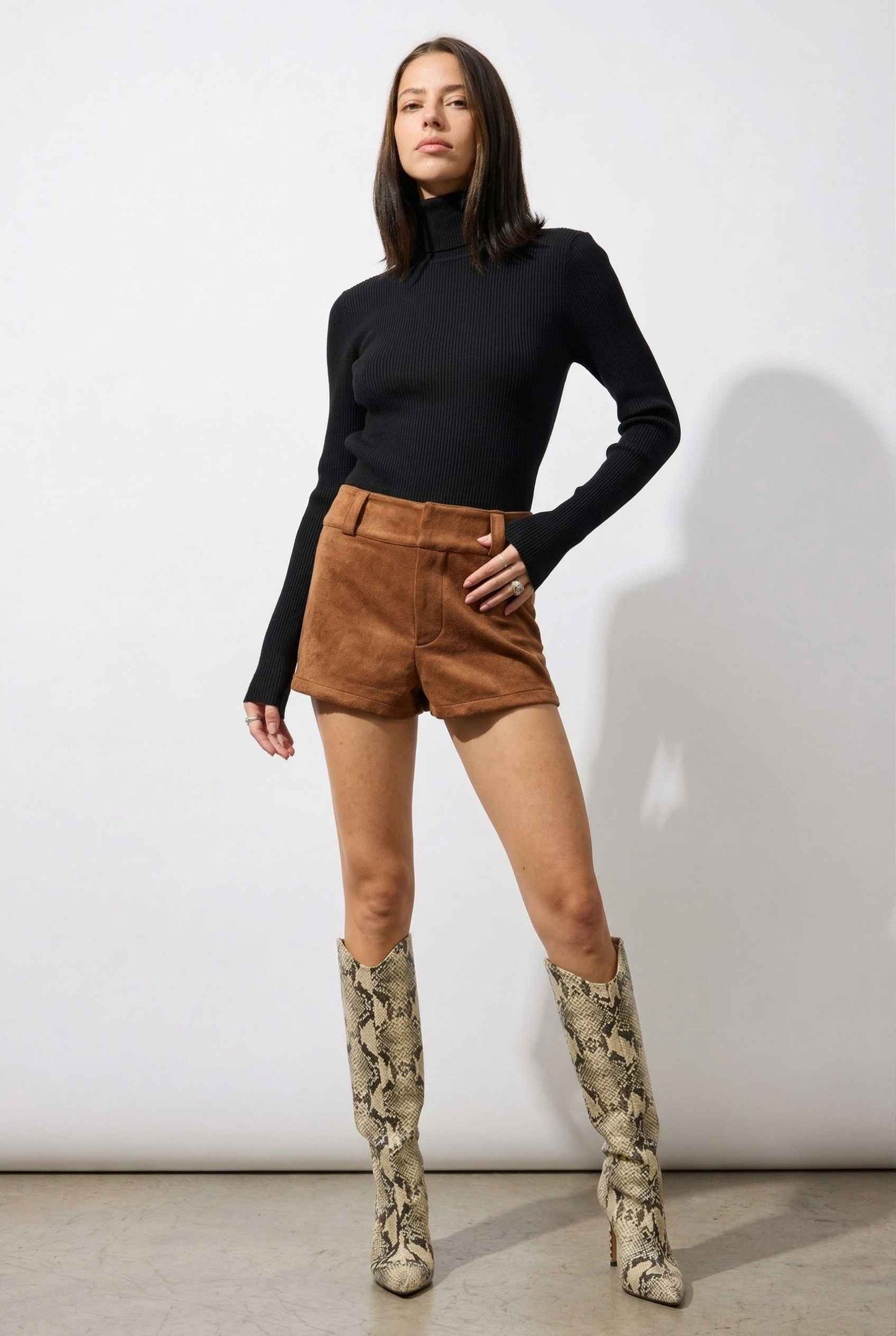 Woman wearing a black turtleneck, Dakota Shorts Brown Faux Suede Low-Rise Mini Shorts, and patterned knee-high boots against a white background