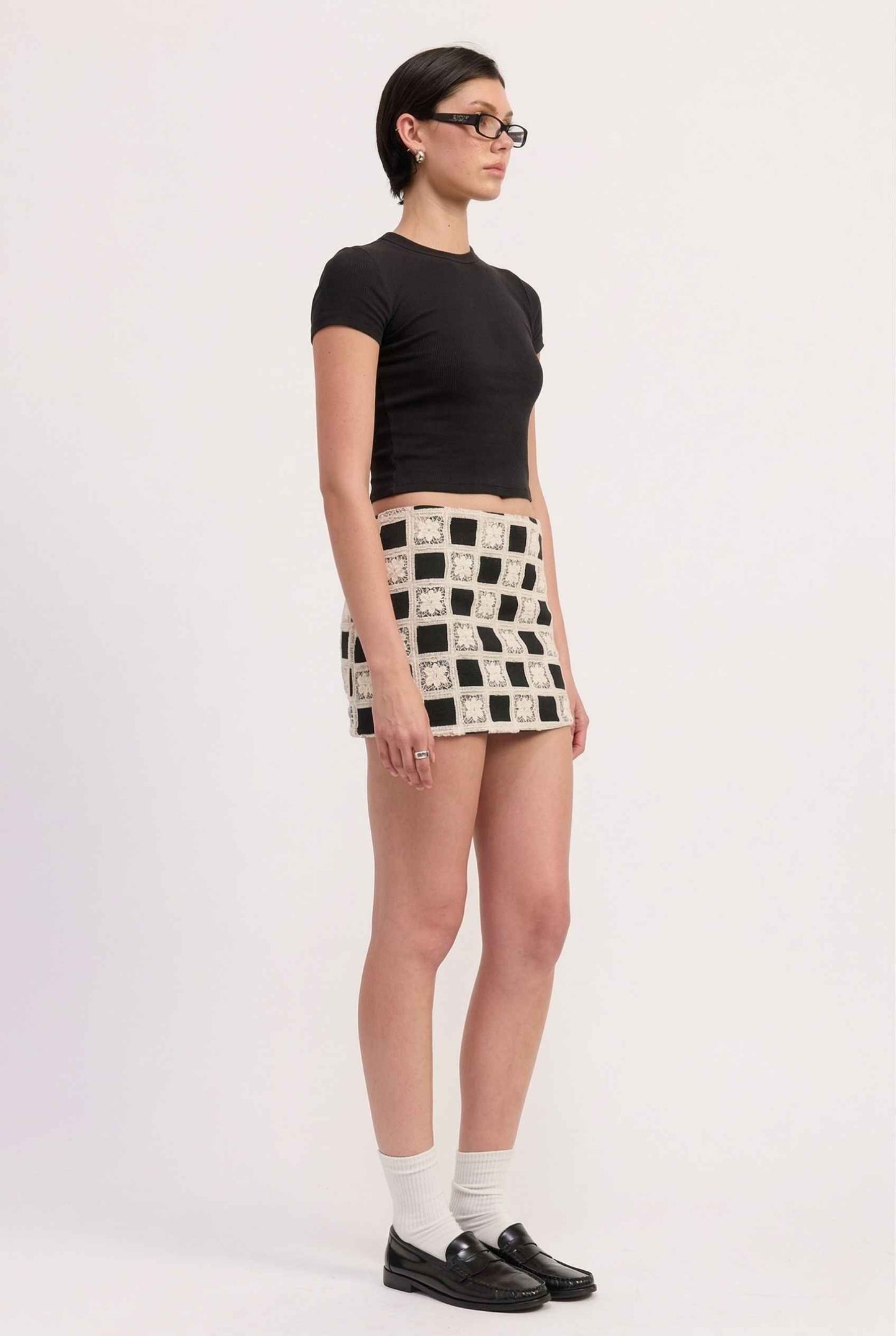 Model wearing Daphne Skirt in Black Modern Jacquard Checkerboard Mini Skirt with basic top and shiny loafers with tall socks