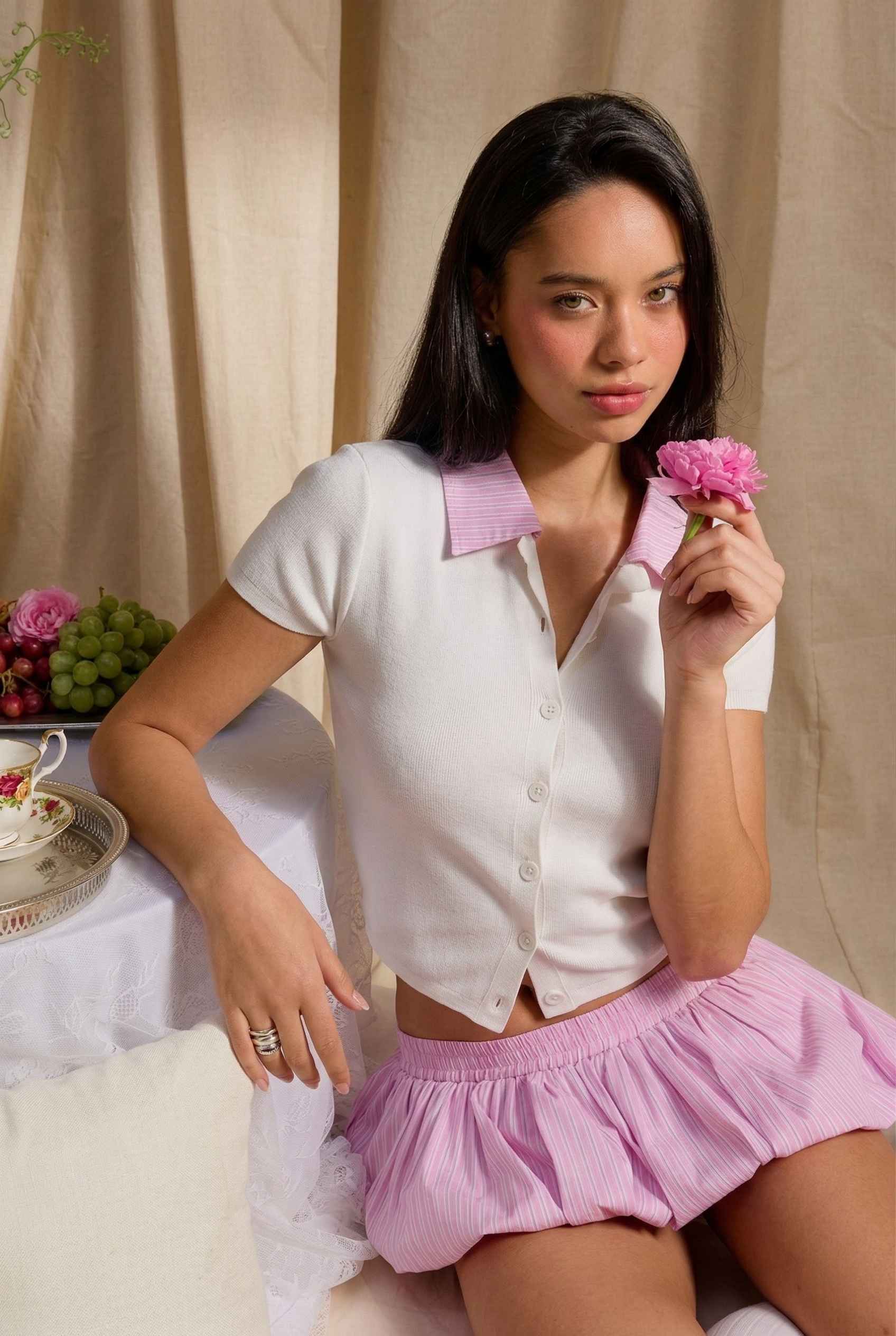 Woman in a Darcy Top White & Pink Contrast Collar Button Down Knit and pink skirt holding a flower, sitting on a couch with a decorative background.
