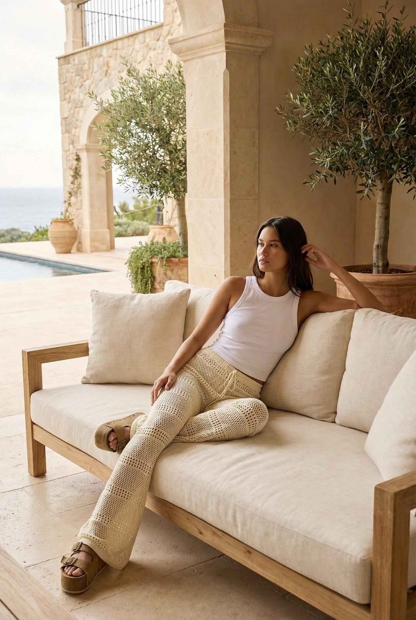 Model wearing Eden Pants in Cream Cotton Crochet Drawstring Beach Trousers with basic tank top and platform sandals sitting on luxury patio by pool