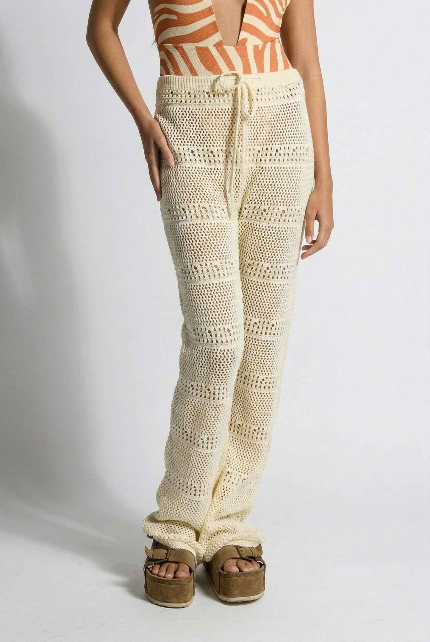 Model wearing Eden Pants in Cream Cotton Crochet Drawstring Beach Trousers with swimsuit and platform sandals on a plain background