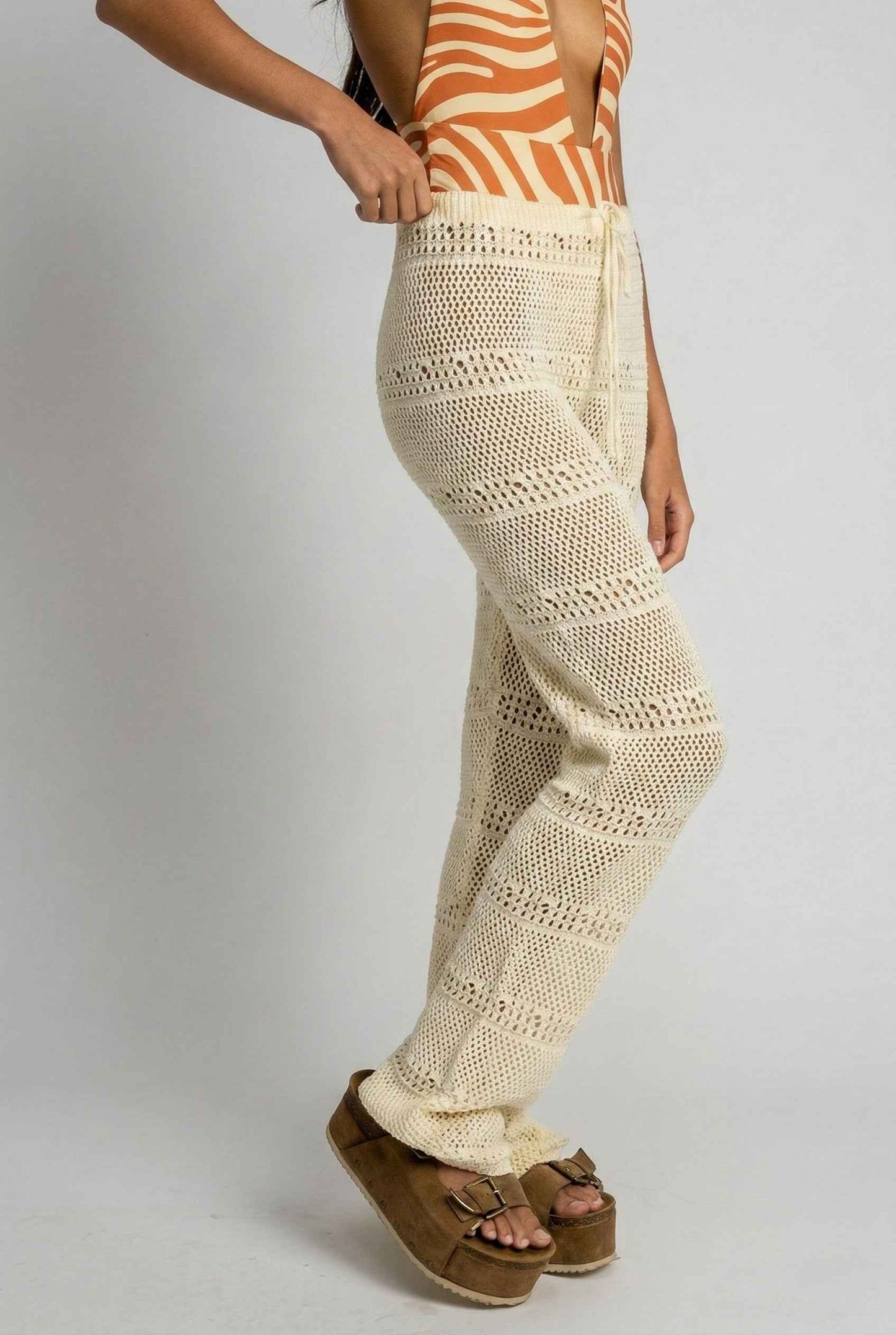 Model wearing Eden Pants in Cream Cotton Crochet Drawstring Beach Trousers with swimsuit and platform sandals on a plain background
