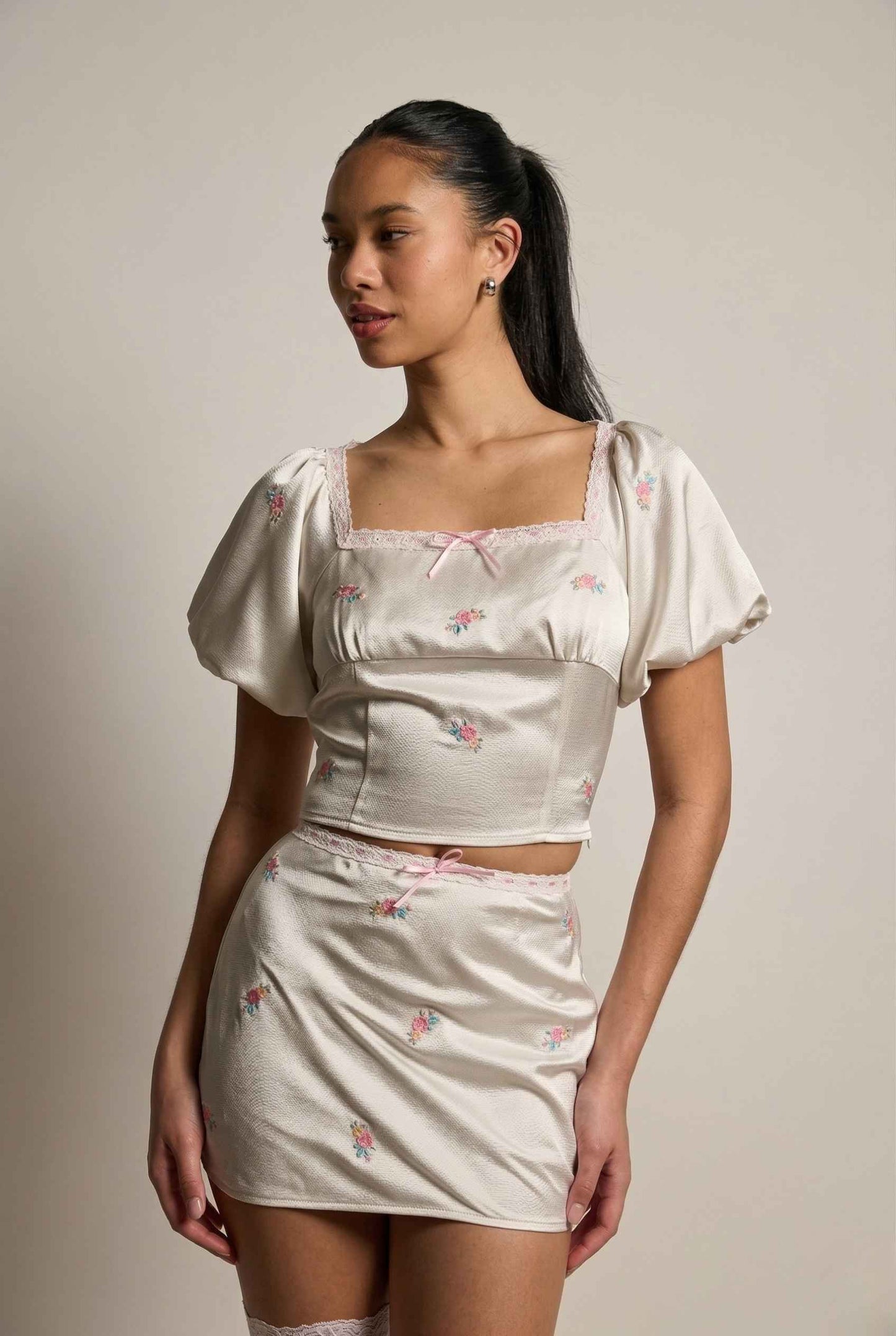 Woman wearing a matching top and Elara Skirt White Satin Mini Skirt with Dainty Pink Florals with floral patterns on a plain background