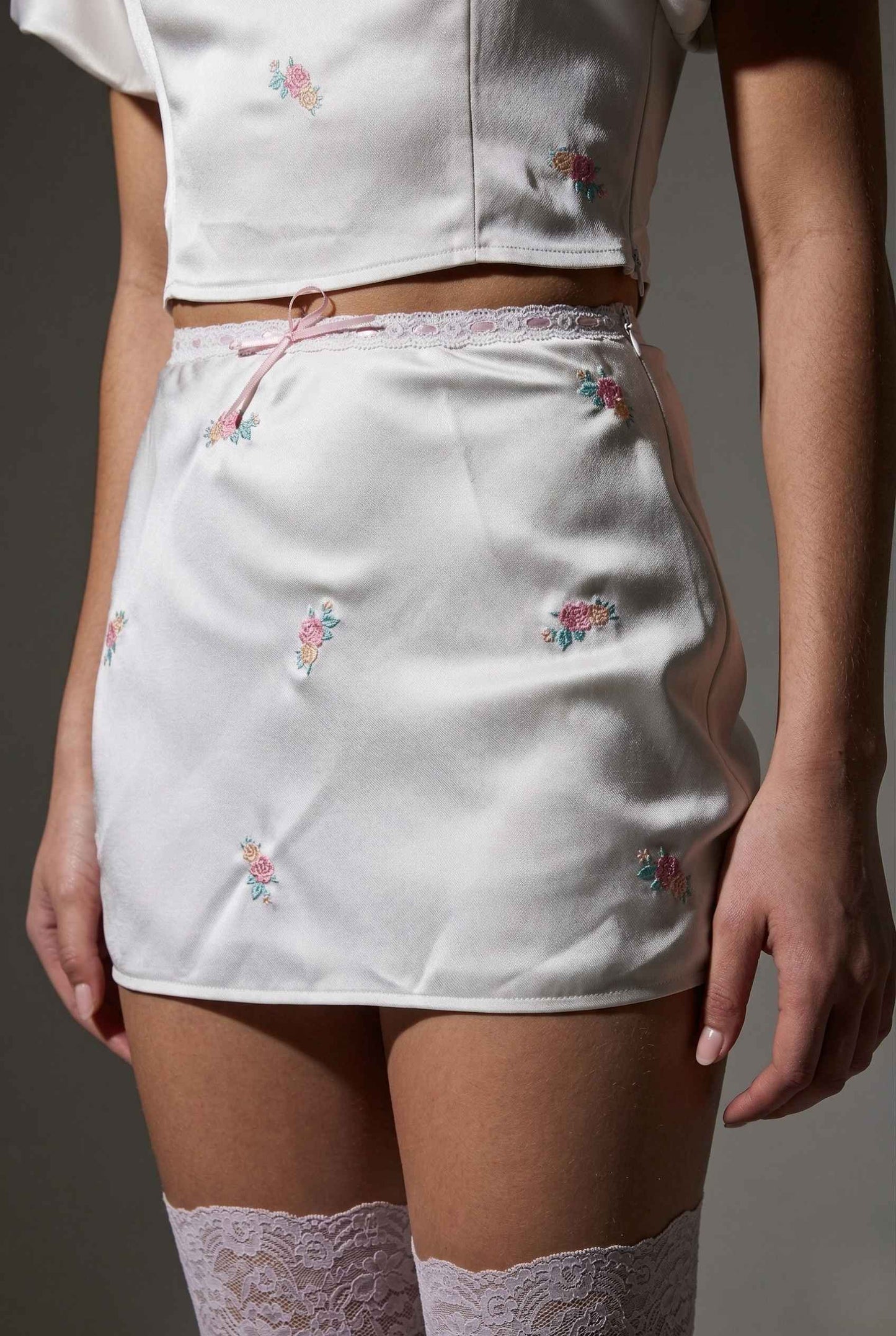 Elara Skirt White Satin Mini Skirt with Dainty Pink Florals with floral embroidery worn by a person on a gray background