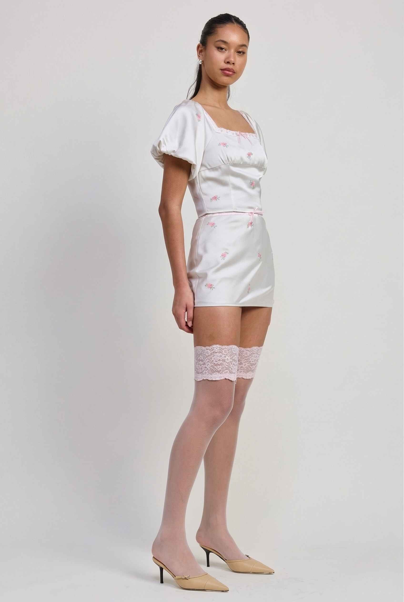 Woman wearing a Elara Skirt White Satin Mini Skirt with Dainty Pink Florals with sheer stockings on a plain background