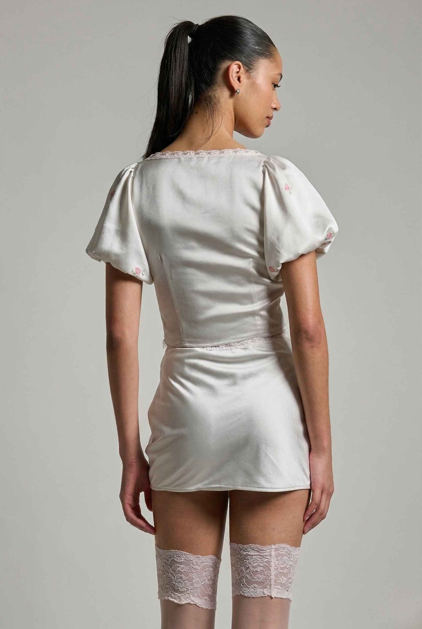 Woman wearing a Elara Skirt White Satin Mini Skirt with Dainty Pink Florals with puffed sleeves against a gray background