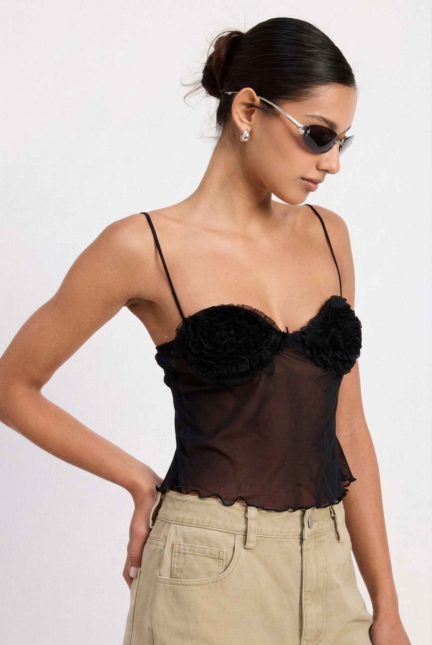 Woman wearing a Elara Top Black Sheer Mesh Camisole with 3D Floral Cups and beige pants against a  background