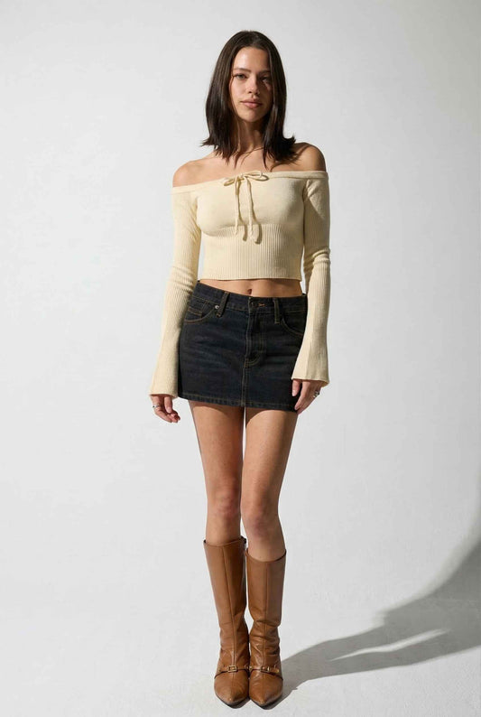 Model wearing Elara Top in Ivory Ribbed Knit Off-Shoulder Tie-Neck Top paired with mini skirt and tall boots on a plain background