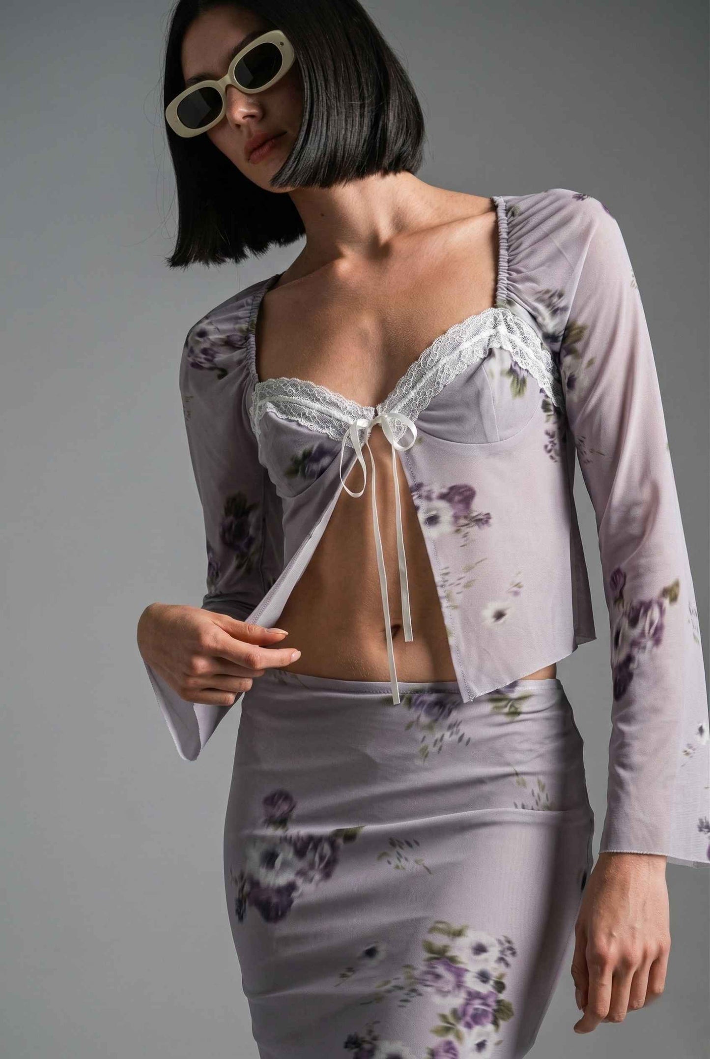 Woman wearing a Elara Top Lavender Floral Lace Trim Fly-Away Cardigan with a sheer overlay against a gray background