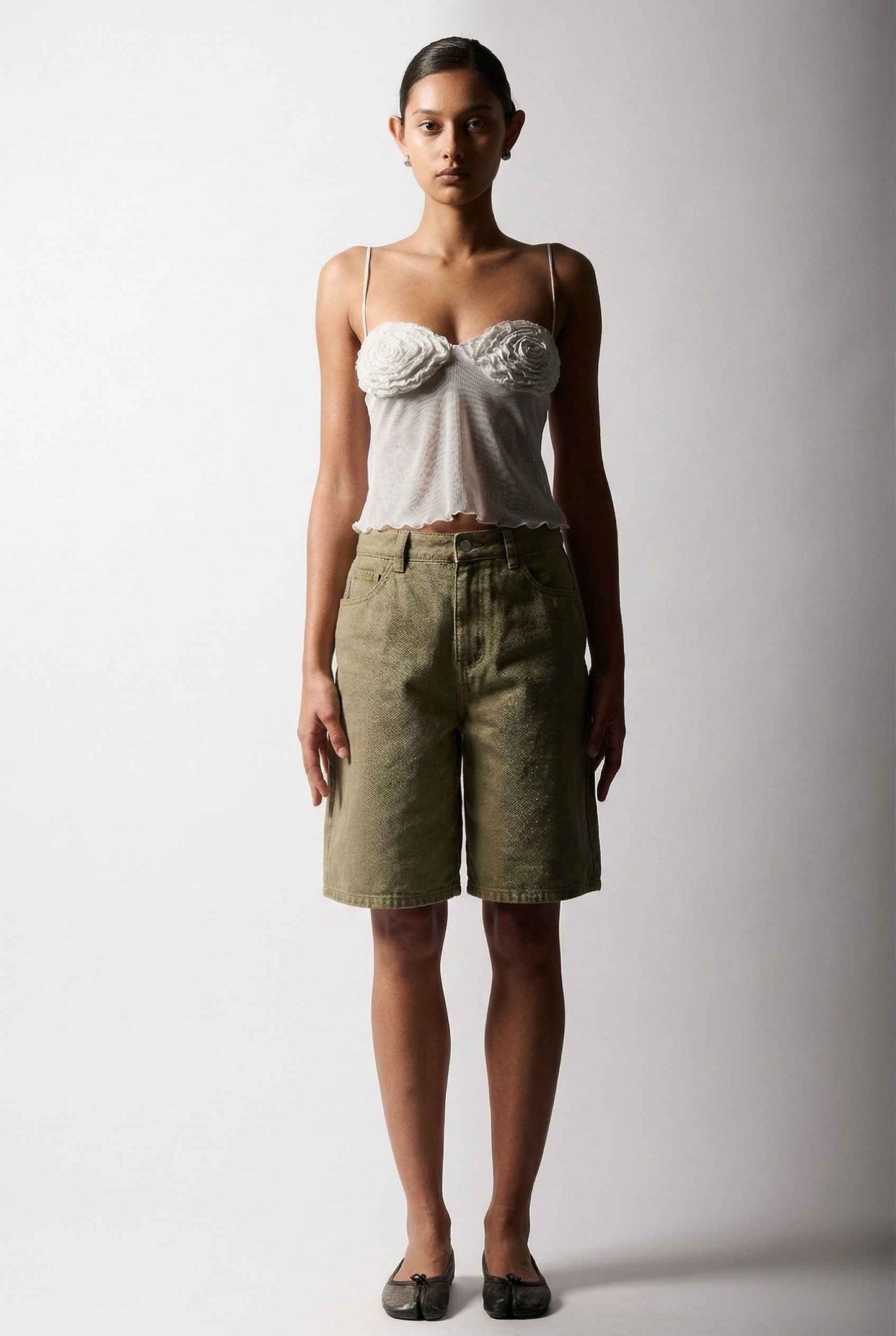 Woman wearing a Elara Top White Sheer Mesh Camisole with 3D Floral Cups and green shorts against a plain background