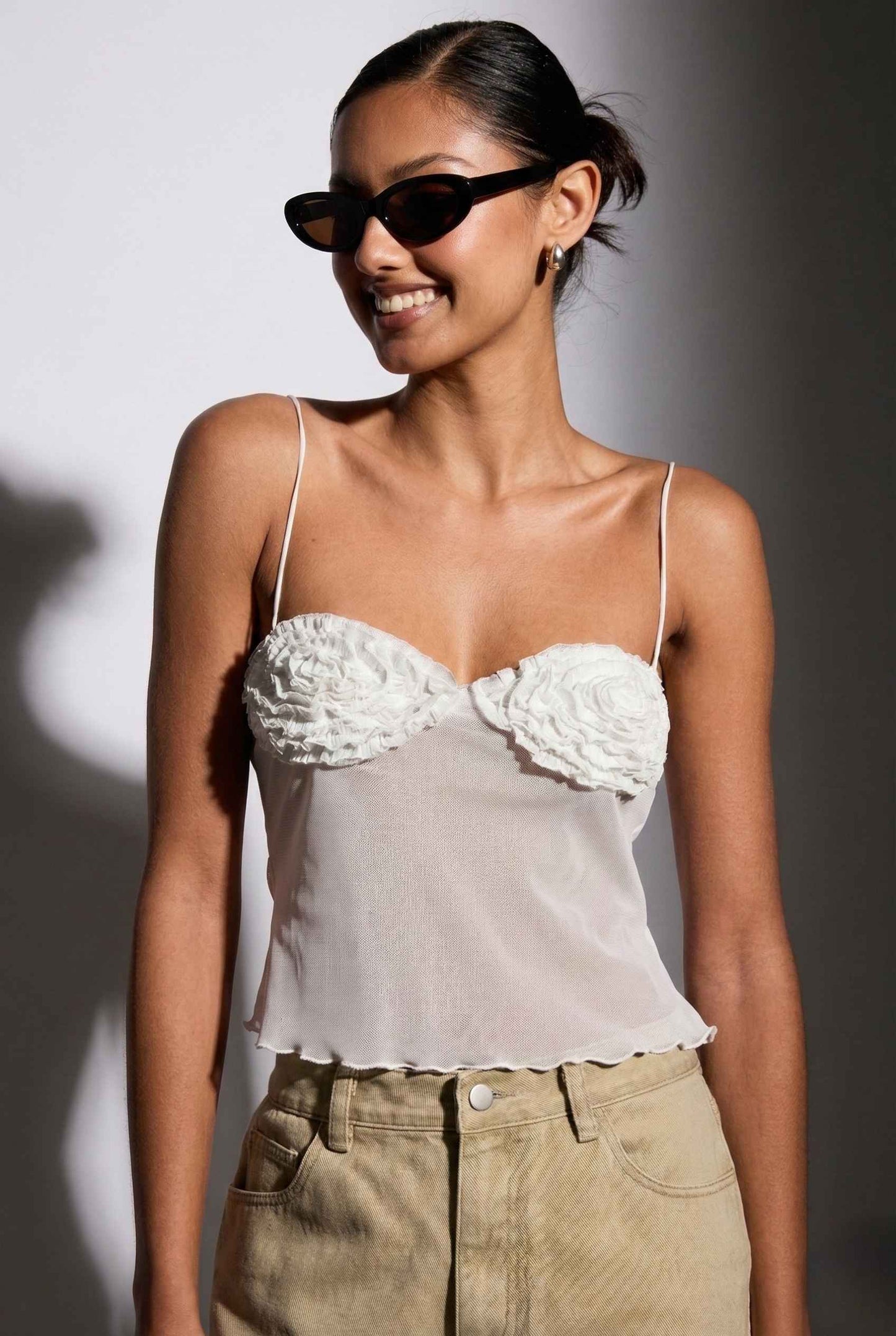 Woman wearing a Elara Top White Sheer Mesh Camisole with 3D Floral Cups and beige pants with sunglasses against a neutral background