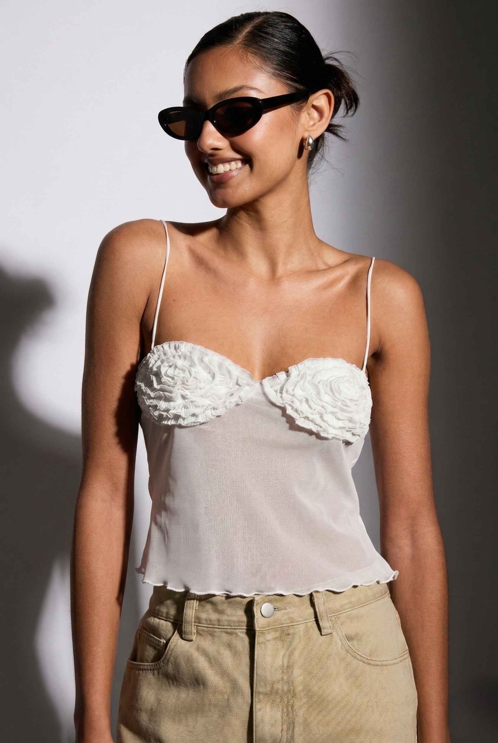 Woman wearing a Elara Top White Sheer Mesh Camisole with 3D Floral Cups and beige pants with sunglasses against a neutral background