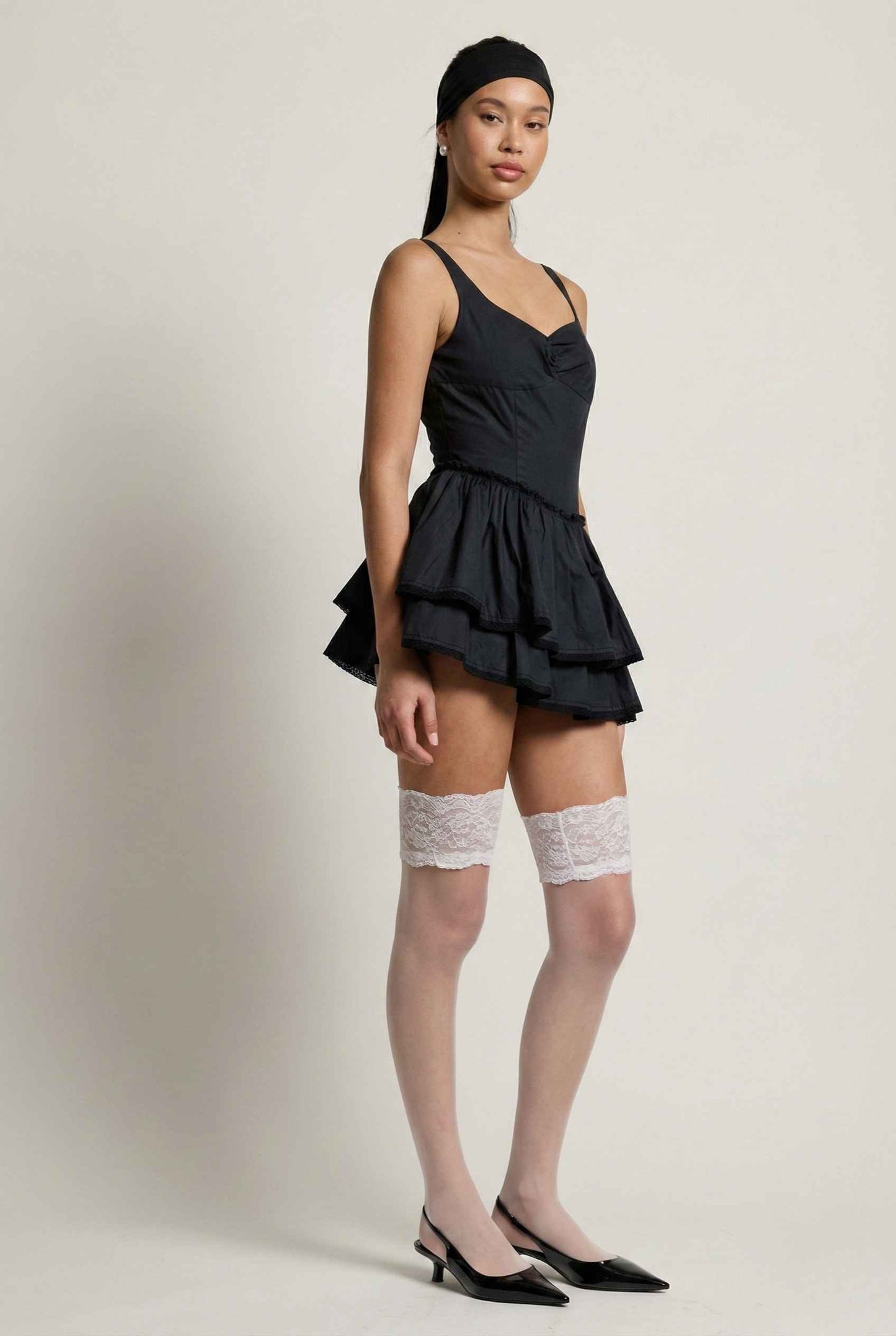 Woman wearing a Elodie Mini Black Tiered Ruffle Cotton Mini Dress with ruffled layers and knee-high socks on a plain background